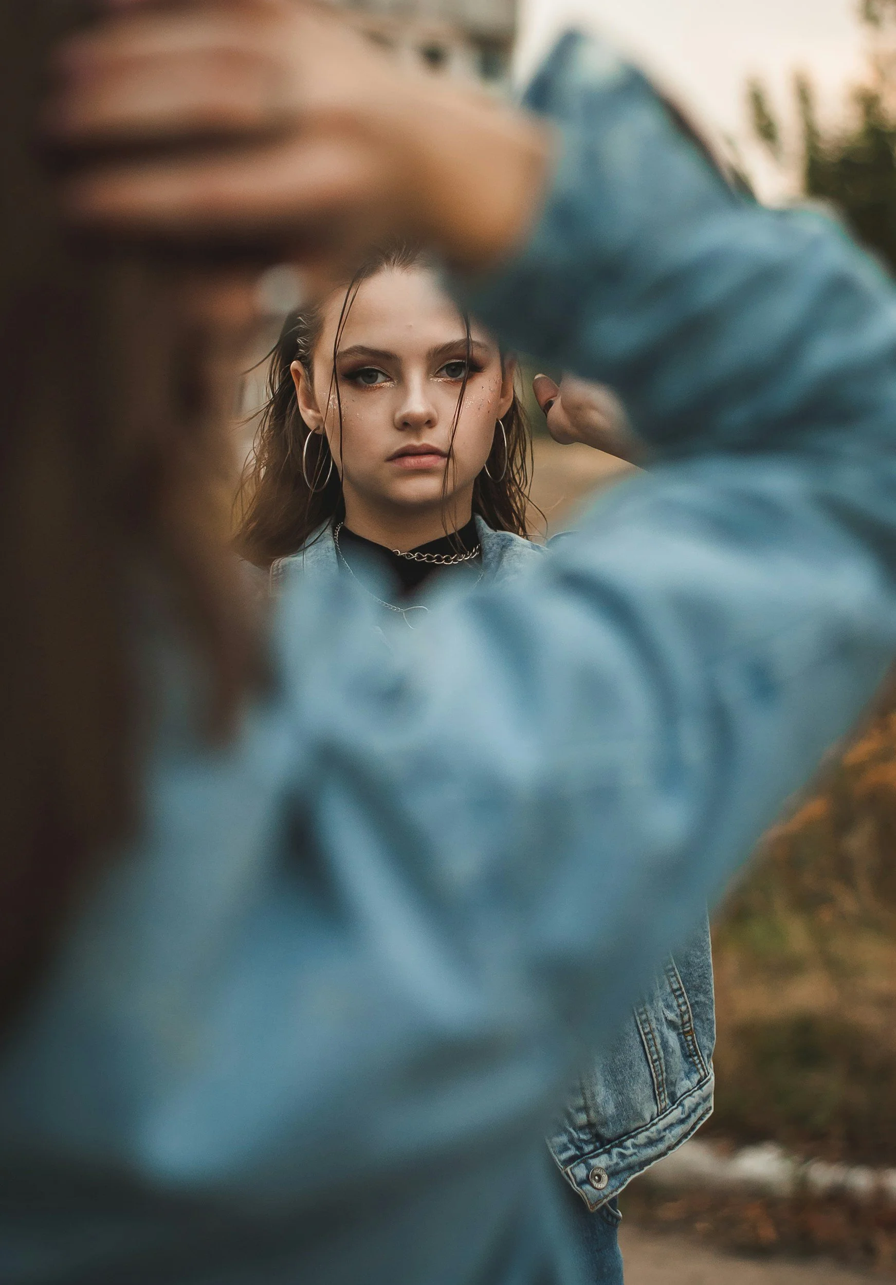 A young woman with brown hair and hoop earrings looking directly at the camera, while someone in a denim jacket adjusts her hair or collar outdoors.