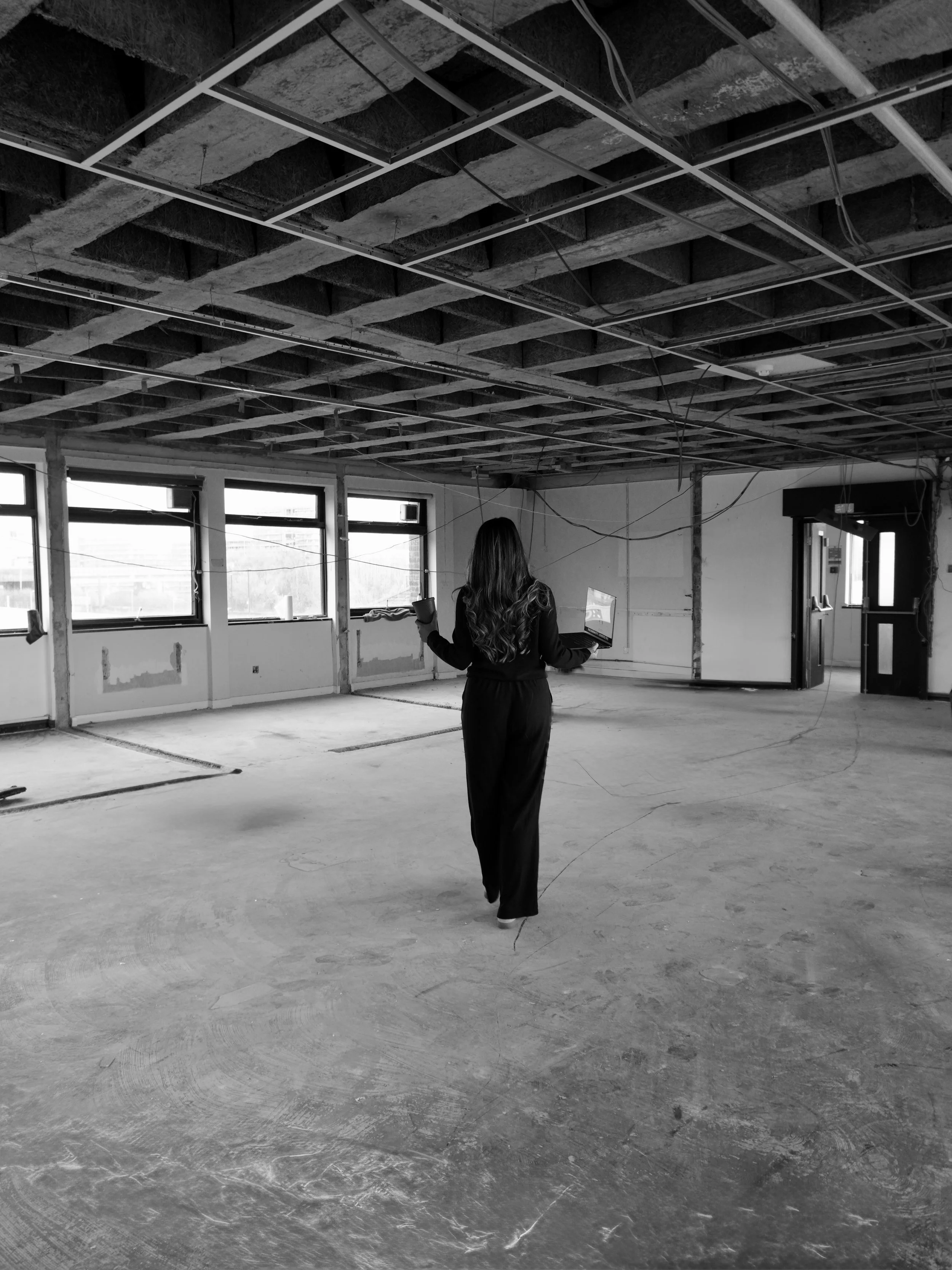 A woman in dark clothing walking through an unfinished room with exposed ceiling framework and large windows, holding a laptop in one hand and a cup in the other.