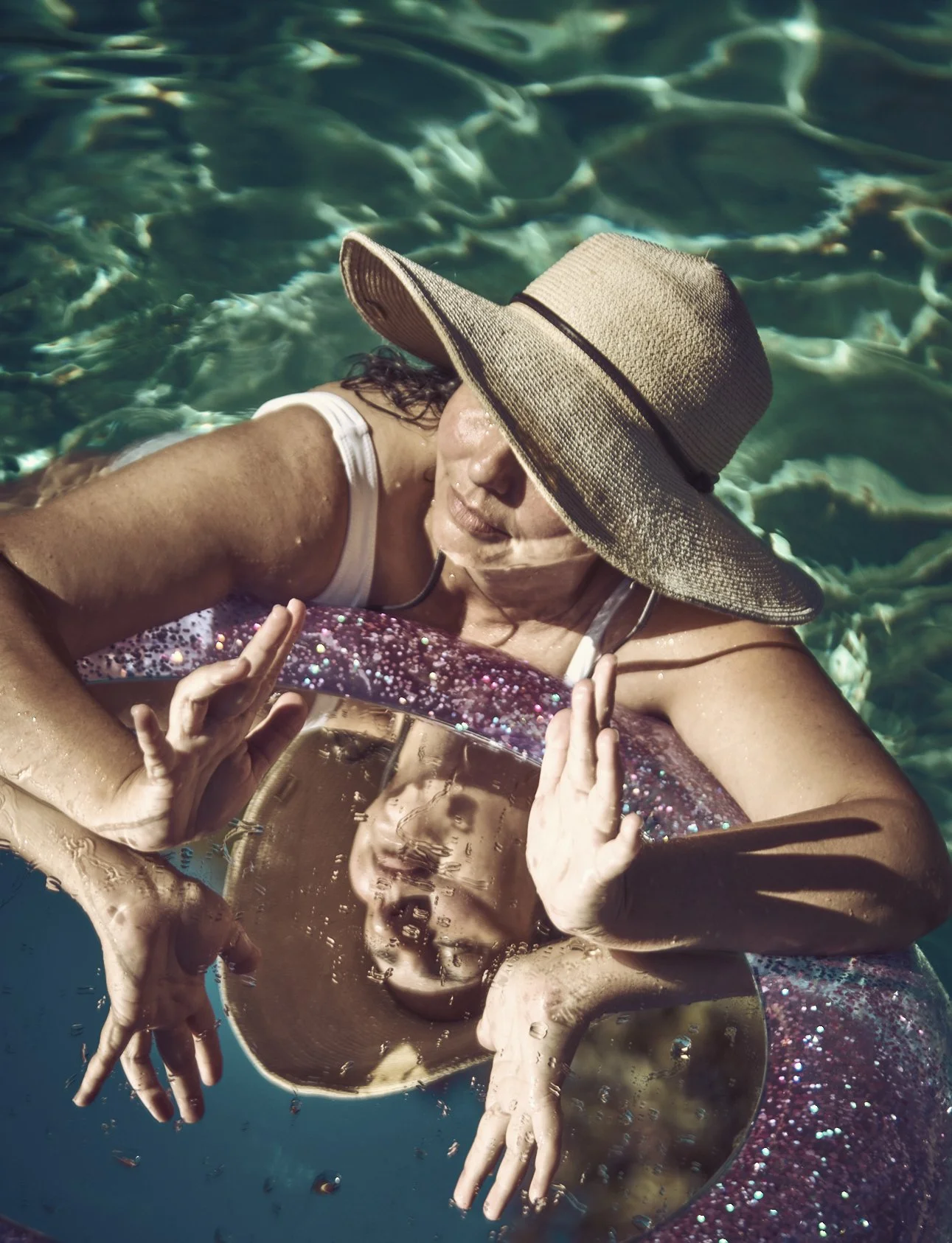 Renata Abbade in a swimsuit and wide-brimmed hat swimming in a pool, holding a mirror, on a glittery pink inner tube. Photo by Chris Clinton.