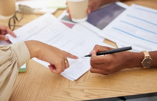 Two people reviewing documents at a wooden table, one pointing and the other holding a pen, with papers and a coffee cup visible.