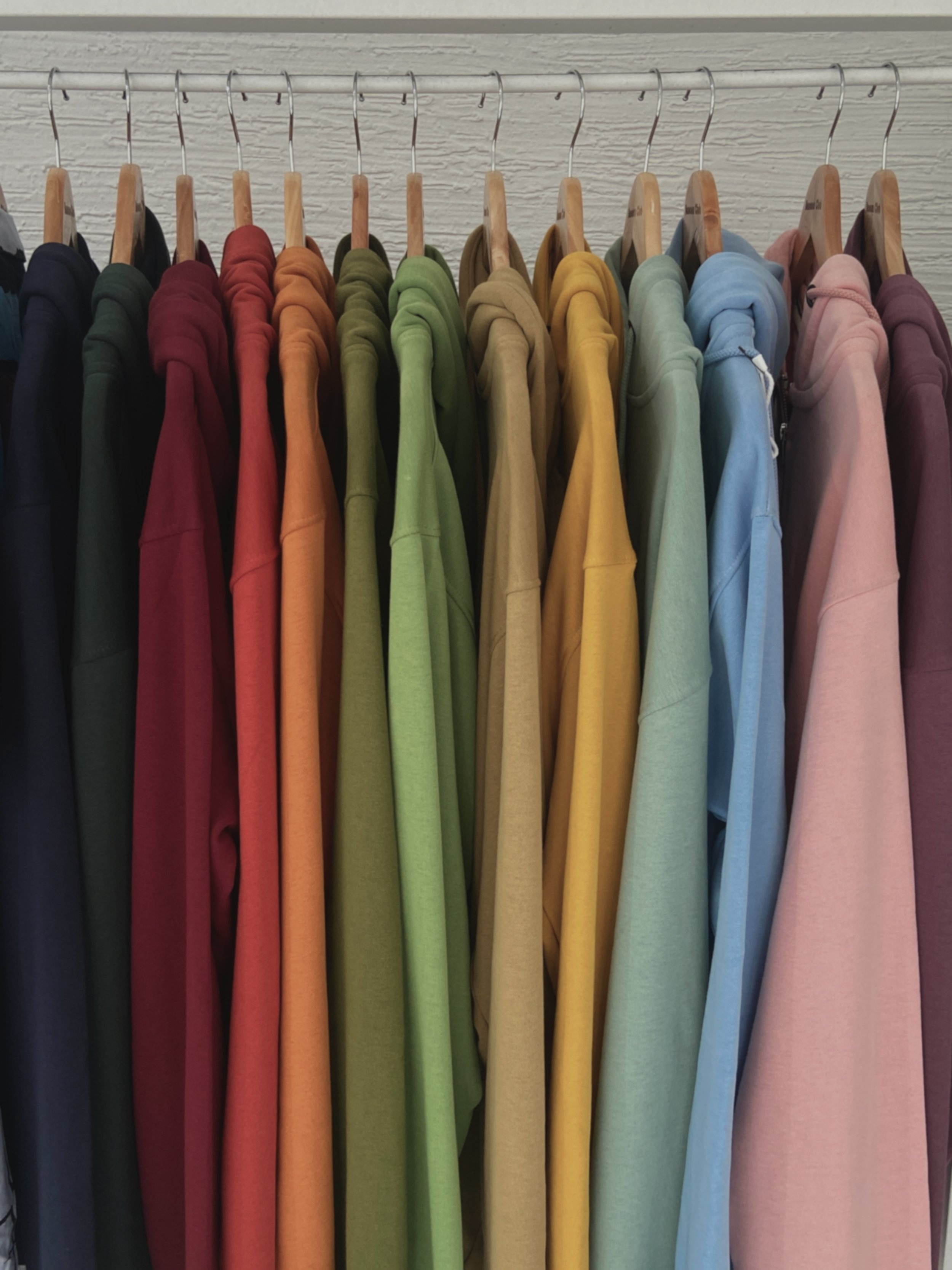 A row of multicolored hoodies hanging on wooden hangers on a metal clothing rack, arranged from dark to light colors.
