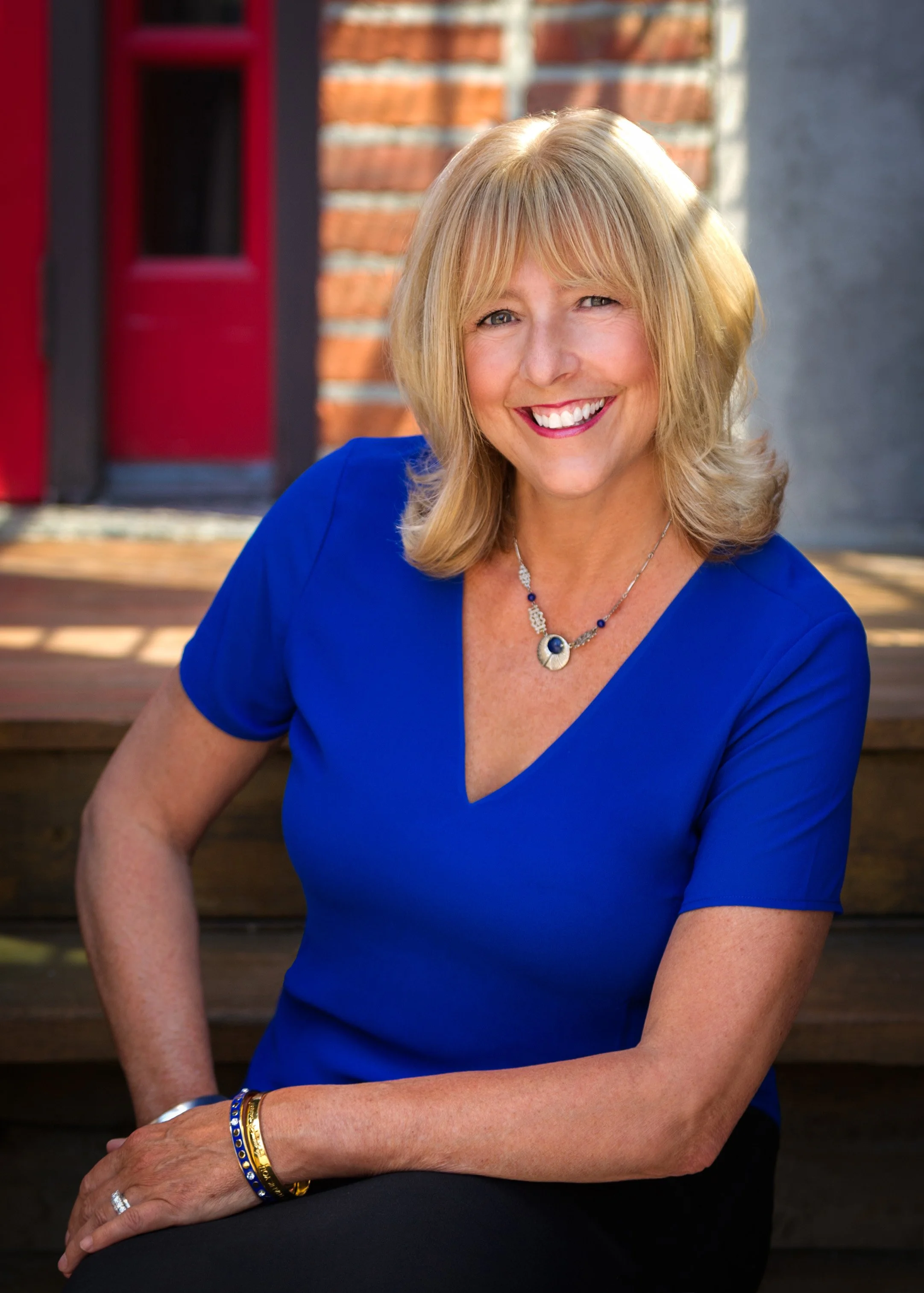 A woman with blonde hair wearing a blue V-neck top, sitting outdoors on wooden steps with a brick wall and red door in the background, smiling.