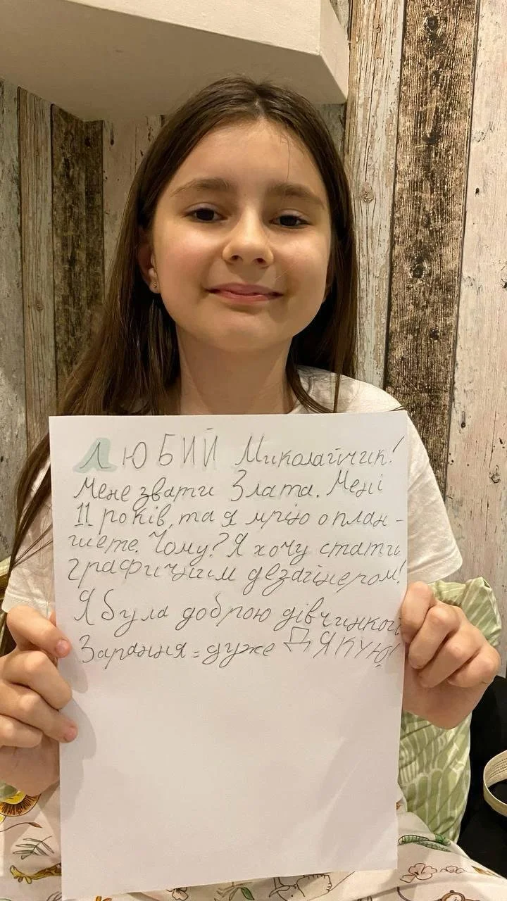 A young girl with long brown hair holding a handwritten sign in Ukrainian. She is smiling slightly and standing in front of a rustic wooden wall.