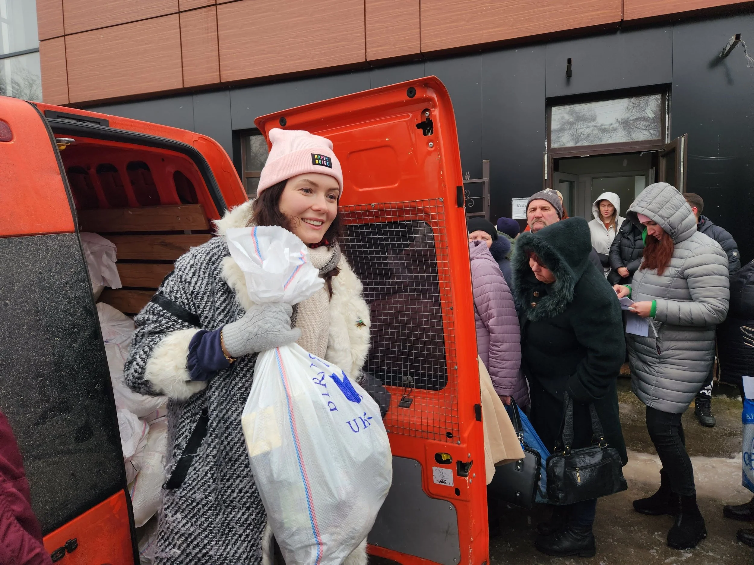 A woman smiling and holding a plastic bag of groceries in front of an orange van, with several people lined up outside a building.
