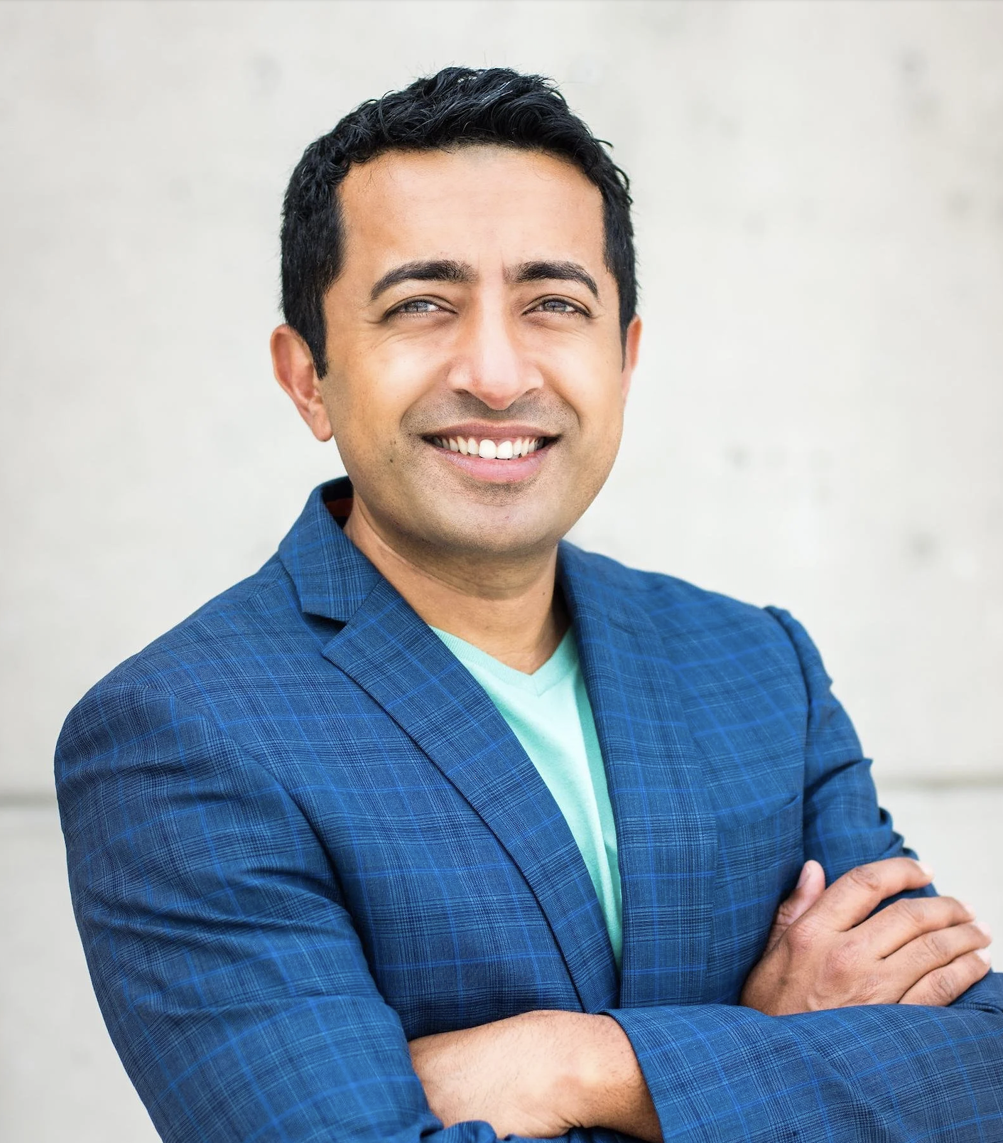 A smiling man with crossed arms, wearing a blue checkered blazer and a light green shirt, standing against a light background.