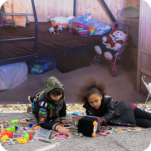 Two children, a boy and a girl, playing on the floor with toys outside their tent. The tent has a bed with stuffed animals, toys, and a Minnie Mouse plush.