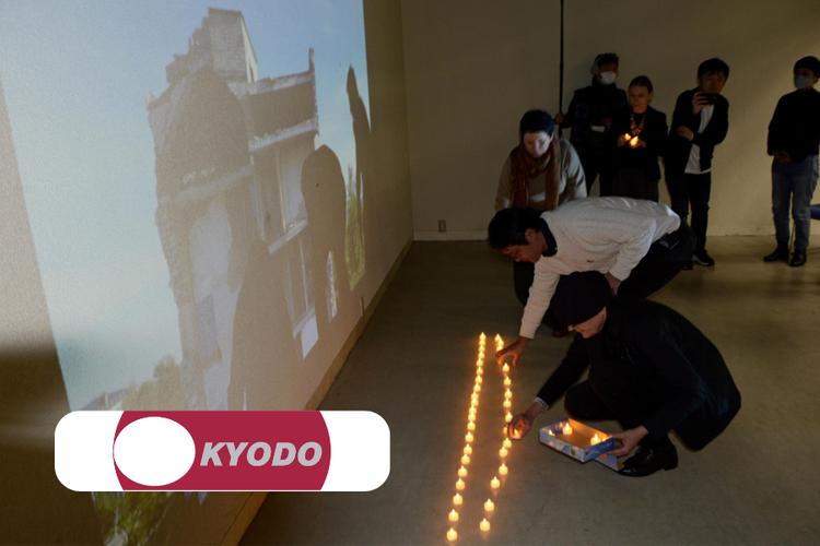 People lighting candles on the floor inside a room to commemorate or honor an event, with a projected image of a historical building on the wall.