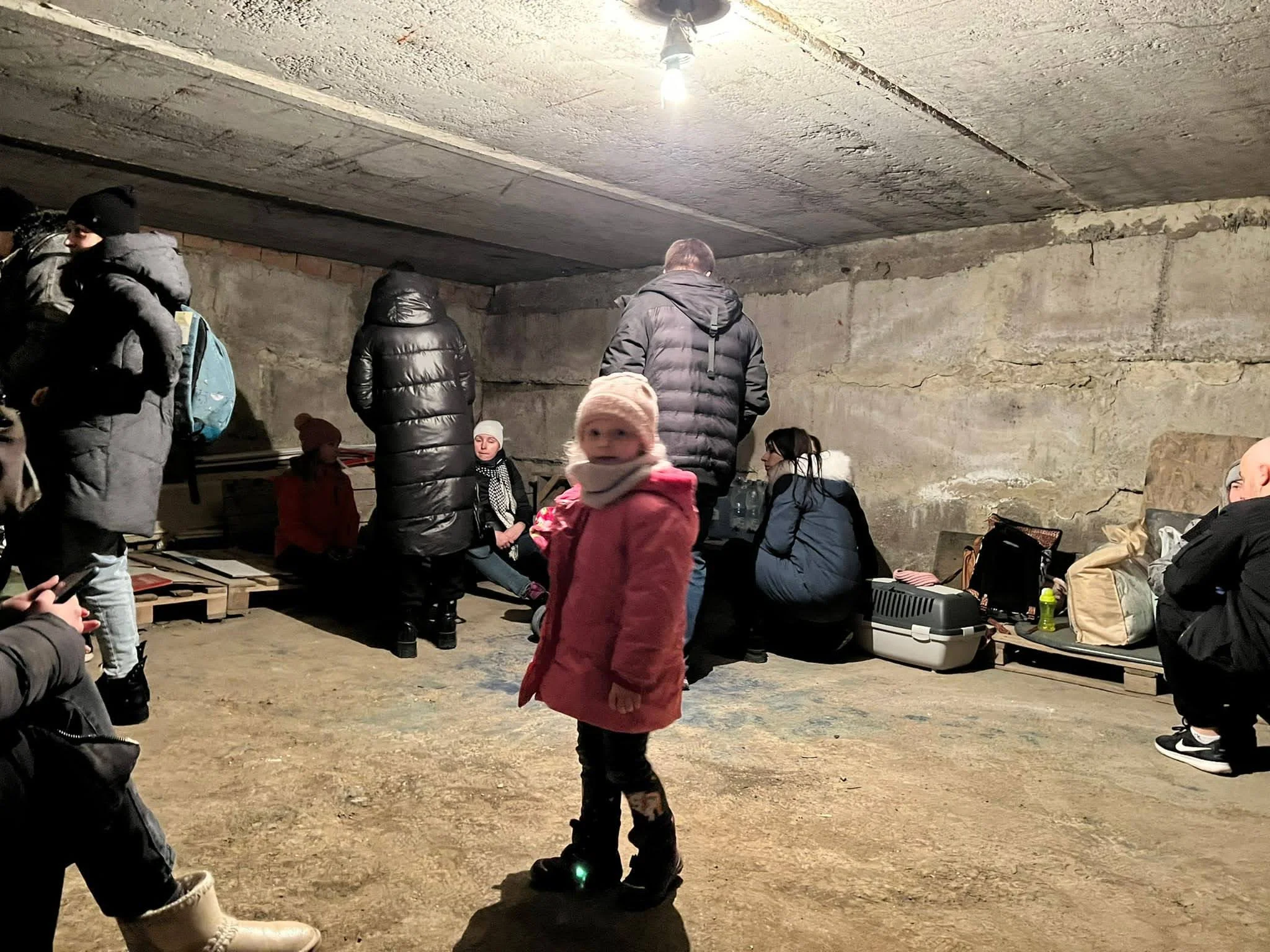 People, including a girl in pink coat and hat, inside a concrete structure with rough walls, some sitting on benches or pallets, others standing or squatting, with bags and supplies around.