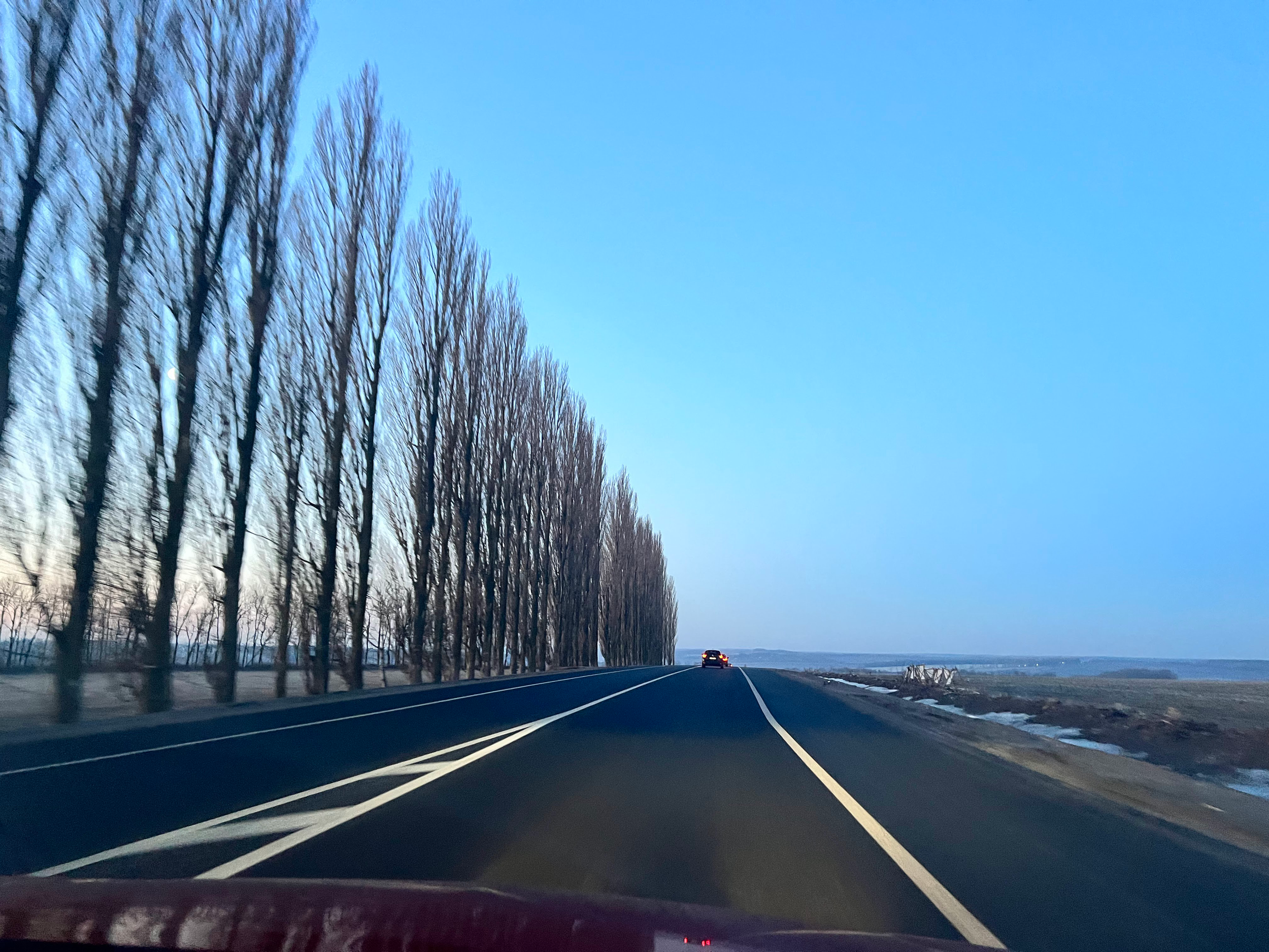 A view from inside a moving vehicle on a highway with leafless trees on the left side and a second vehicle ahead. The sky is clear and blue with some patches of snow on the ground.