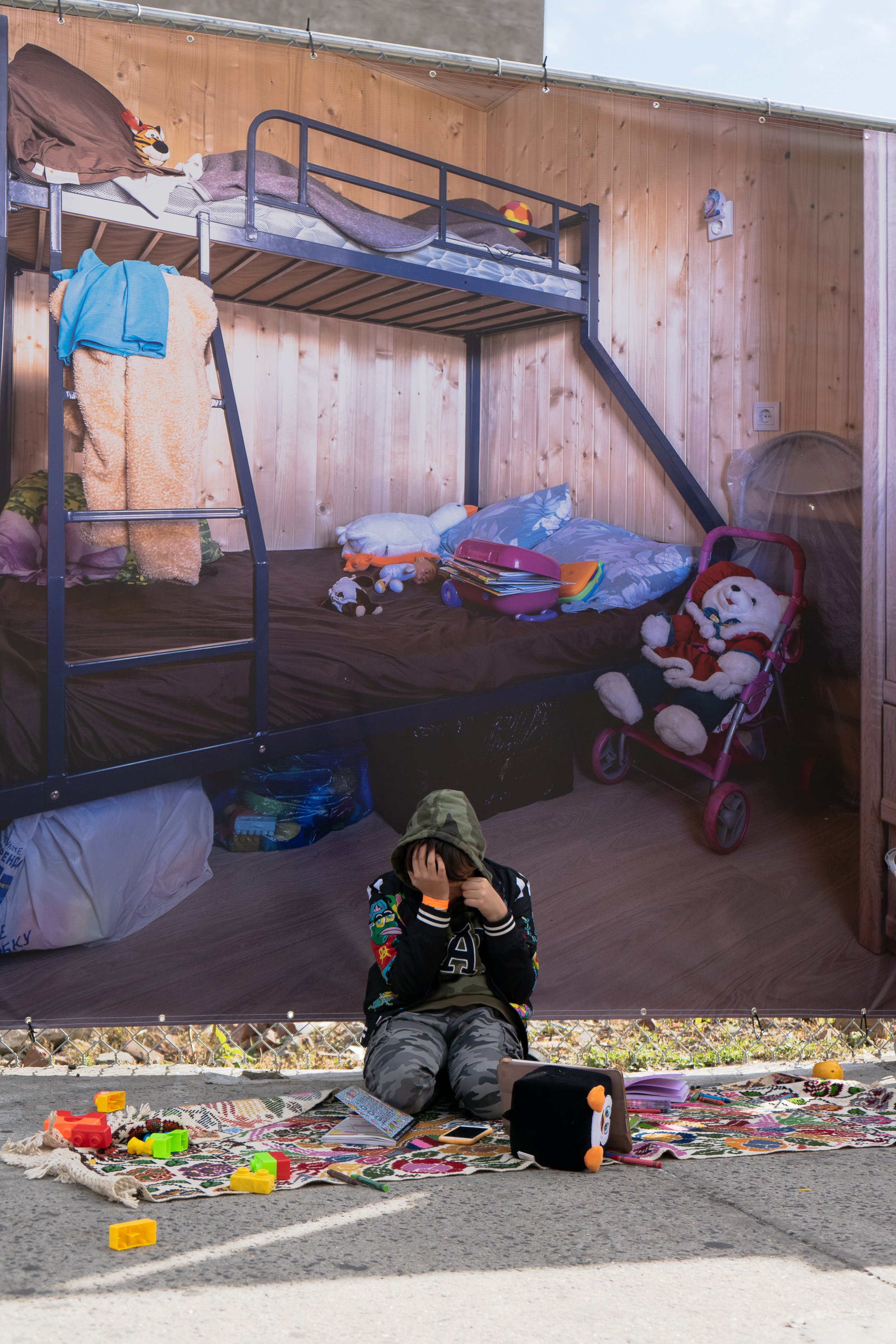 A boy sitting on a colorful rug outside with toys around him, covering his face. Behind a transparent wall, there is a children's bedroom with bunk beds, stuffed animals, and toys.