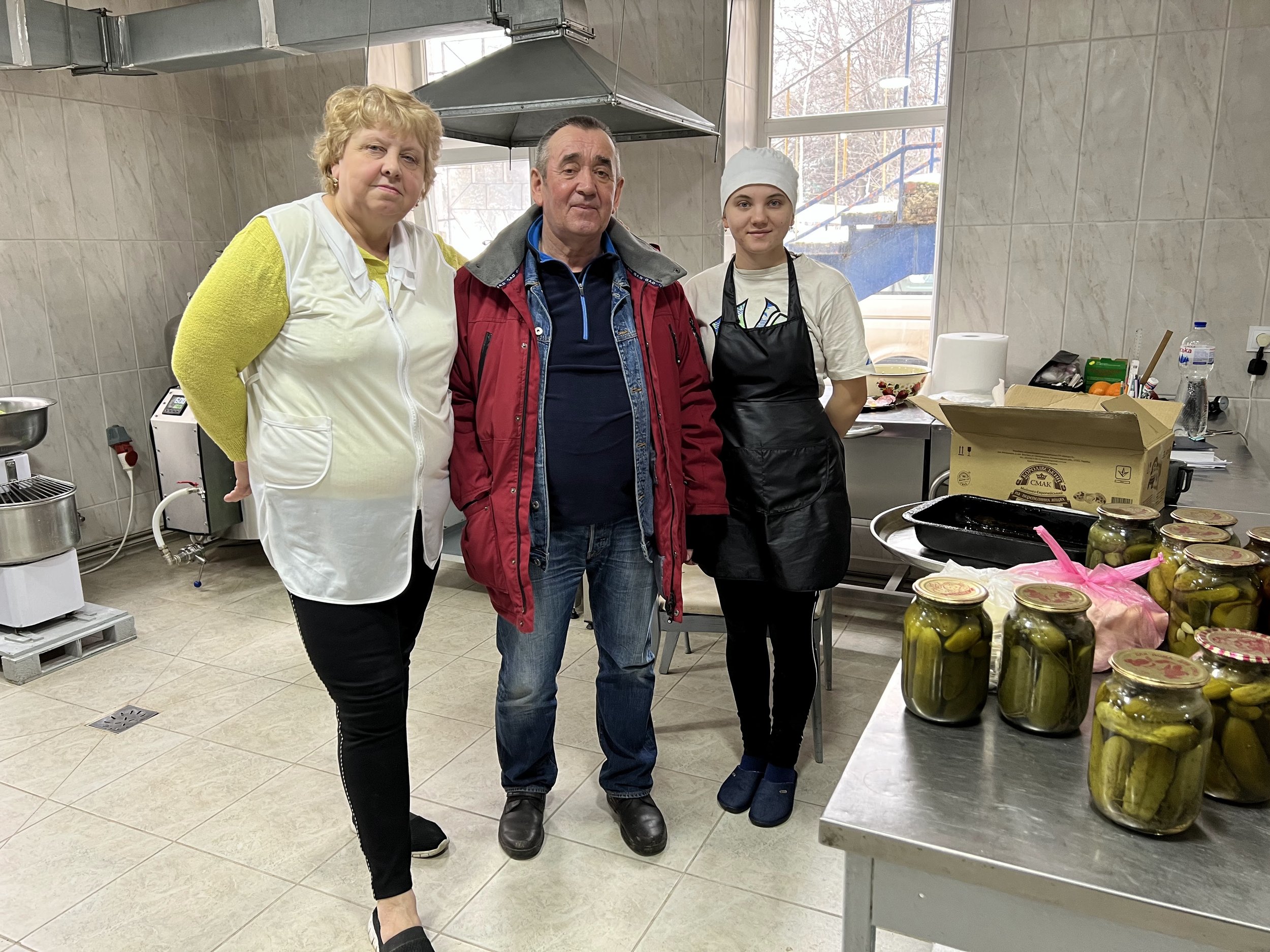 Three people in a kitchen, an older woman and man, and a young woman in a white chef's hat and apron. They are standing behind a table with jars of pickles, with kitchen equipment and window in background.