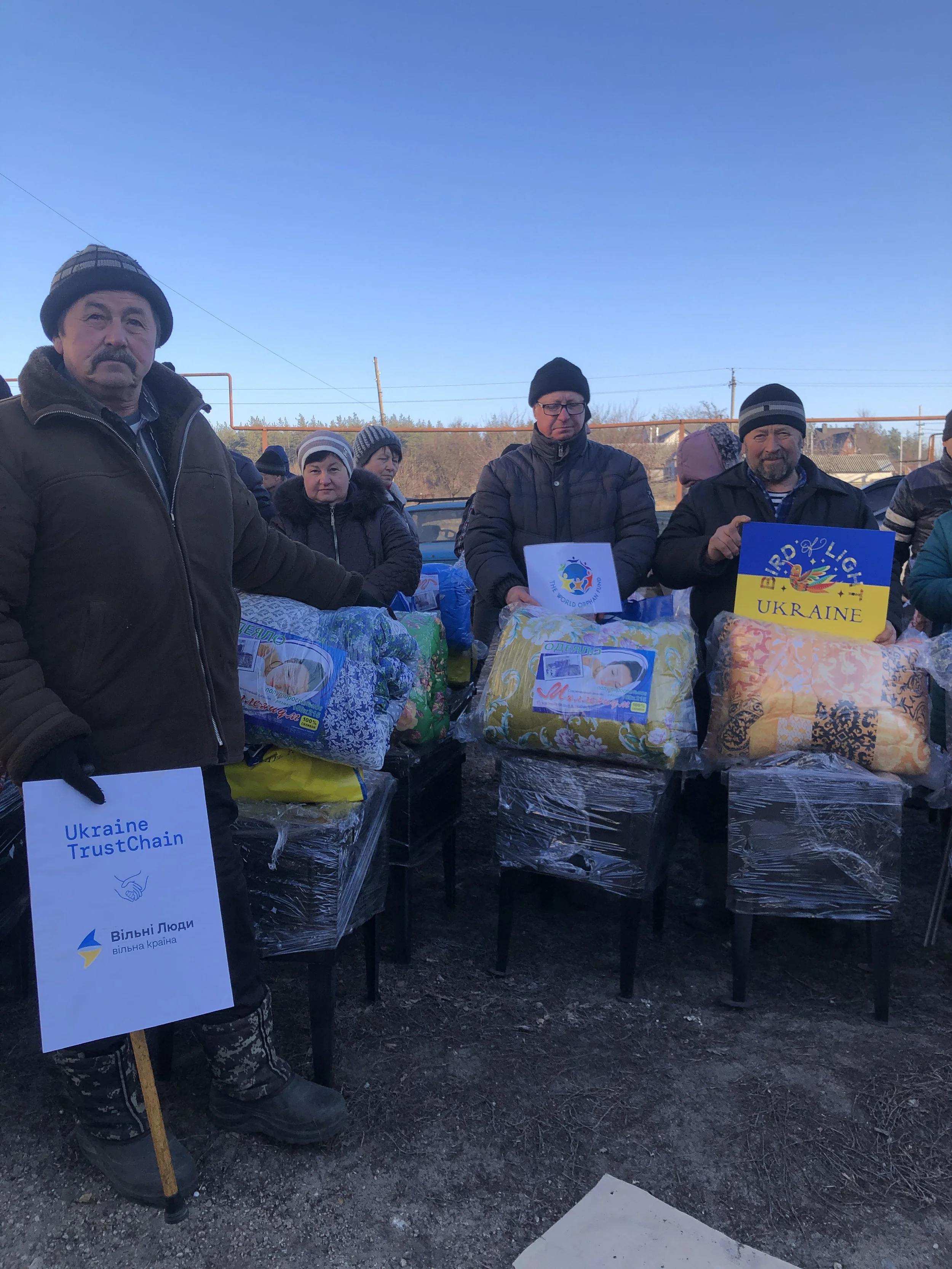 Ukrainian civilians in a grey zone village holding wrapped packages and Ukrainian flags outdoors during daytime.