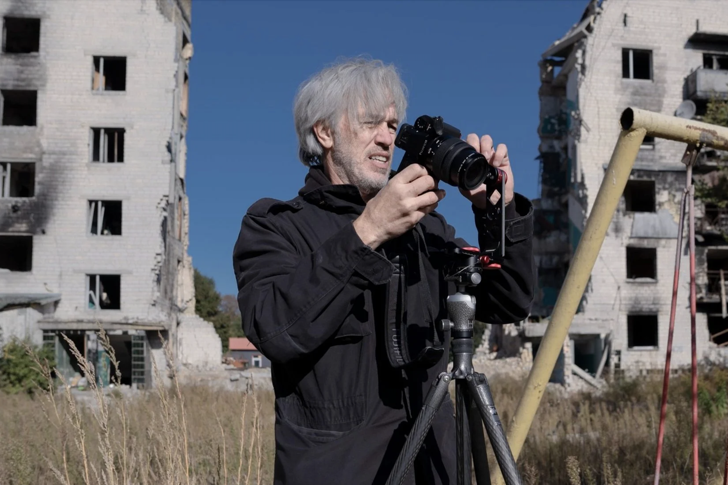 An older man with white hair, wearing a black jacket, is setting up a camera on a tripod. Behind him are damaged buildings with broken windows, in a field with tall grass under a clear blue sky.