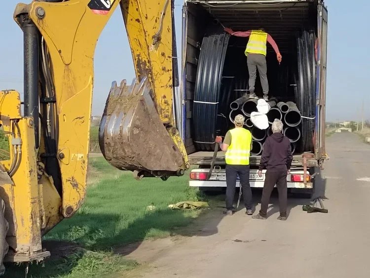 Three UKrainian workers unload pipes from a truck, one standing inside the truck and two on the ground, near a yellow excavator on a road.