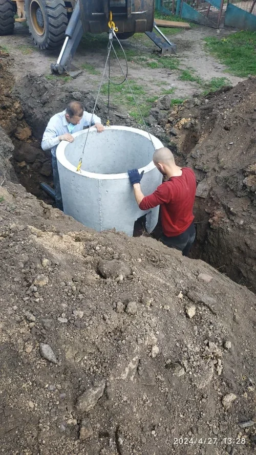 Two Ukrainian men are installing a large concrete pipe into a dug hole in the ground, with a large piece of construction equipment above supporting the pipe.