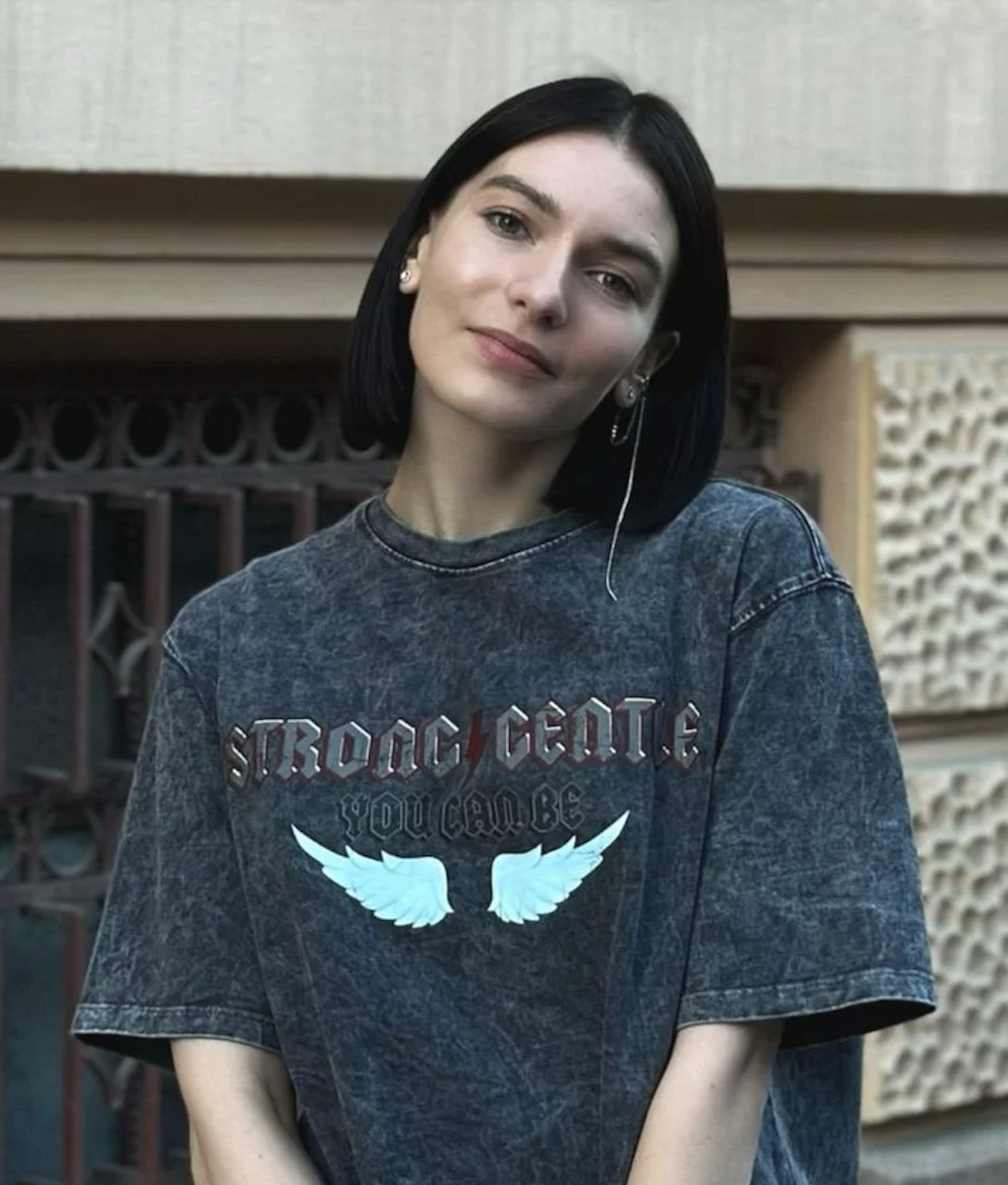 A young woman with black hair and fair skin standing in front of a textured wall, wearing a dark gray T-shirt with angel wings and text that reads "Strong, Gentle, You Can Be." She has a slight smile, wears earrings, and an in-ear headset.