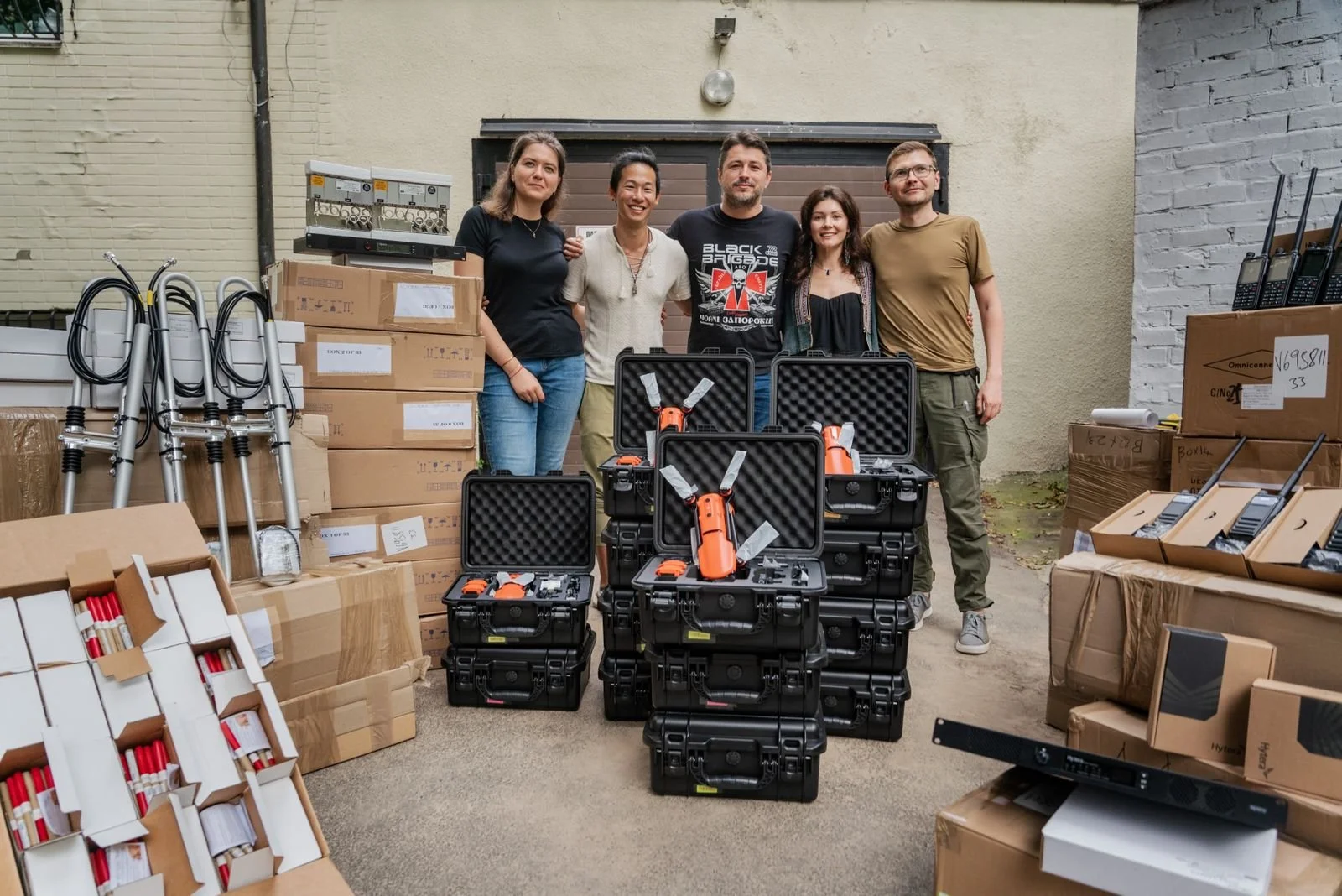 A group of five people standing behind a variety of equipment and supplies, including orange handheld tools in protective cases, boxes of supplies, and crutches, in an outdoor area with a brick and painted wall.