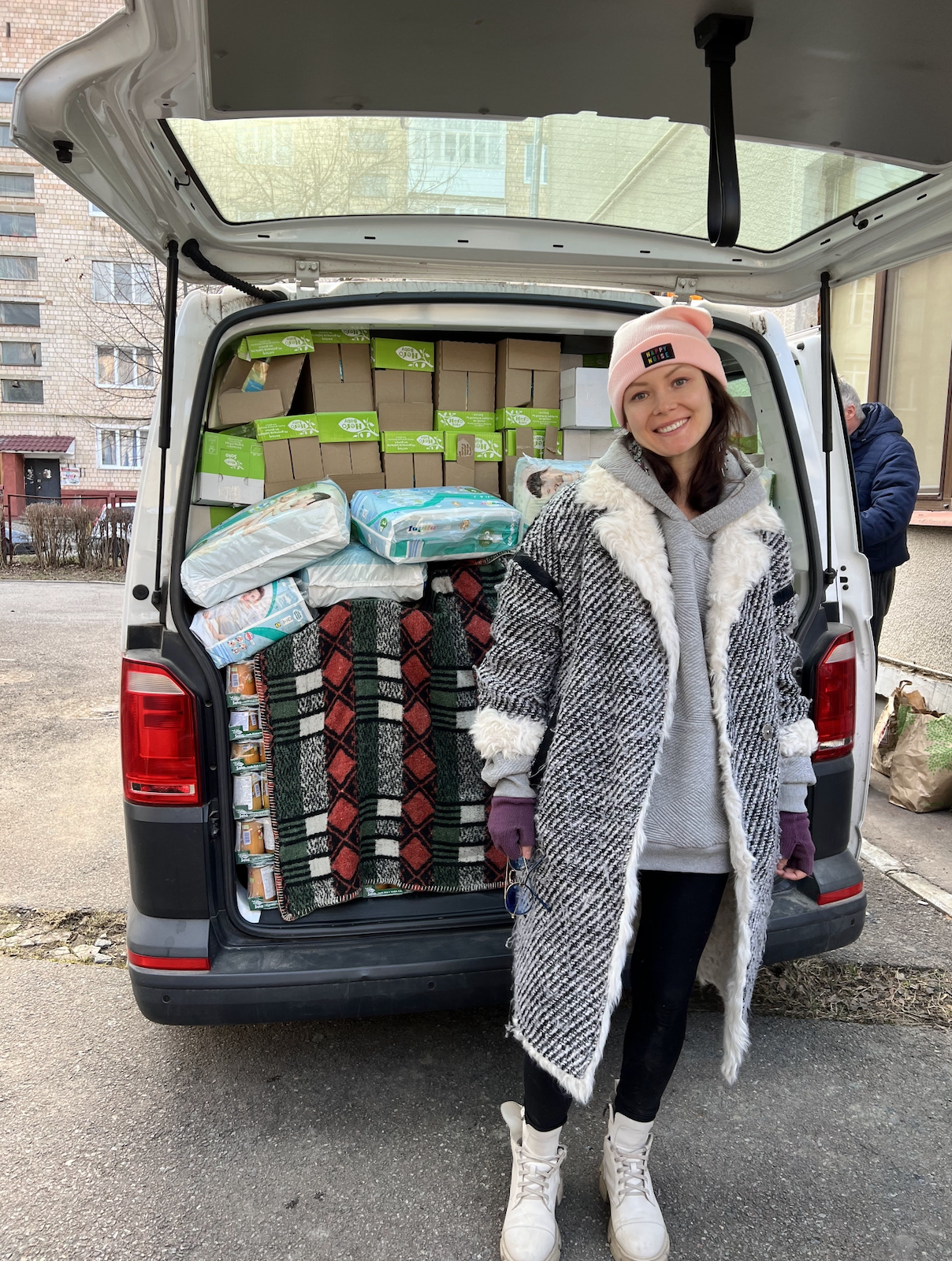 A smiling young woman wearing a pink beanie, gray hoodie, and a long plaid coat standing in front of the open trunk of a van loaded with boxes and packages, outdoors in an urban area.