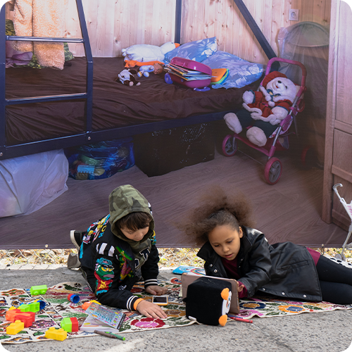 Two children playing on a colorful rug outside a wooden shed, surrounded by toys. The shed has a bed with stuffed animals, pillows, and some books, and a Santa Claus doll is seated in a stroller nearby.