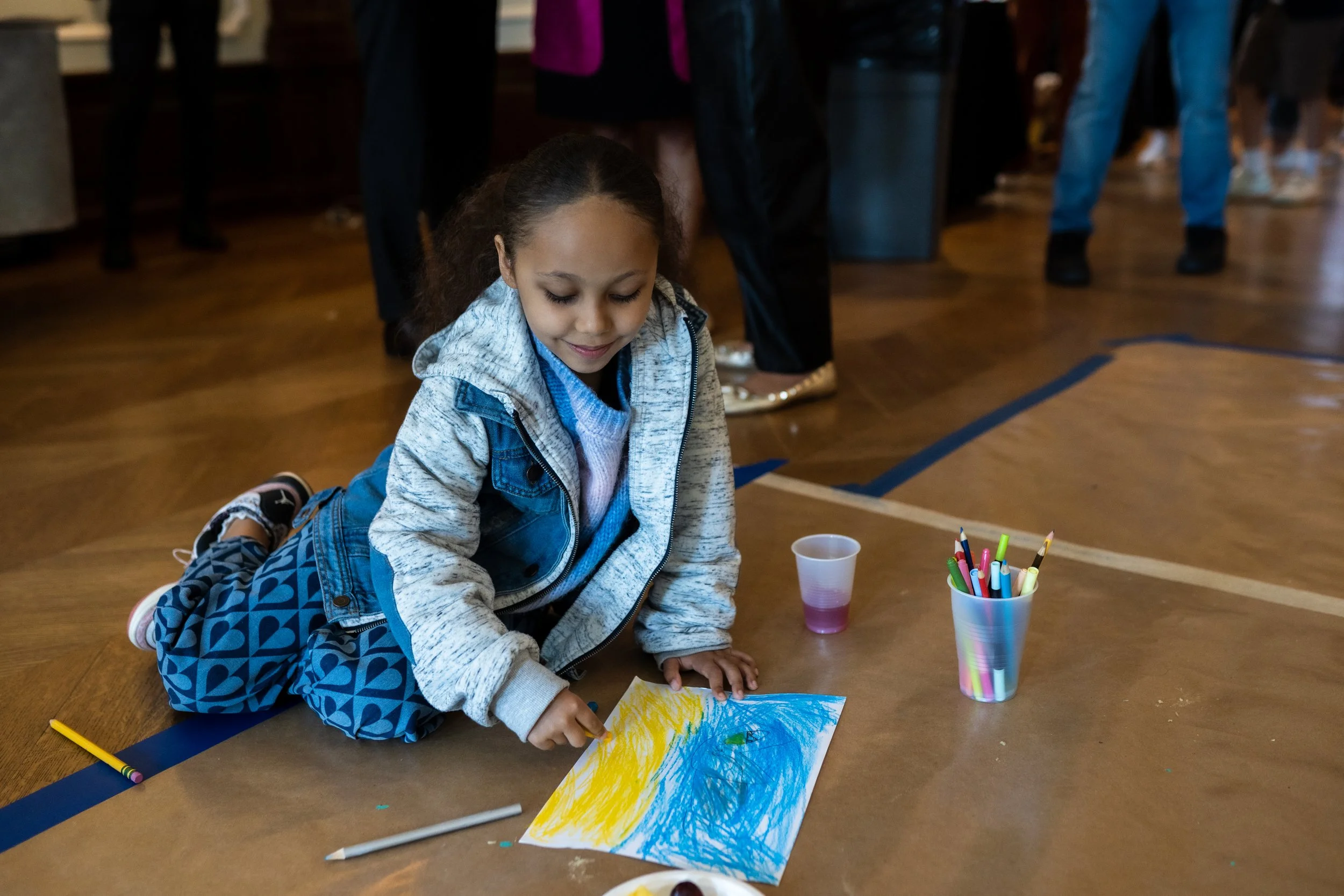 Young girl drawing on a piece of paper using colorful markers, sitting on the floor with art supplies around her.