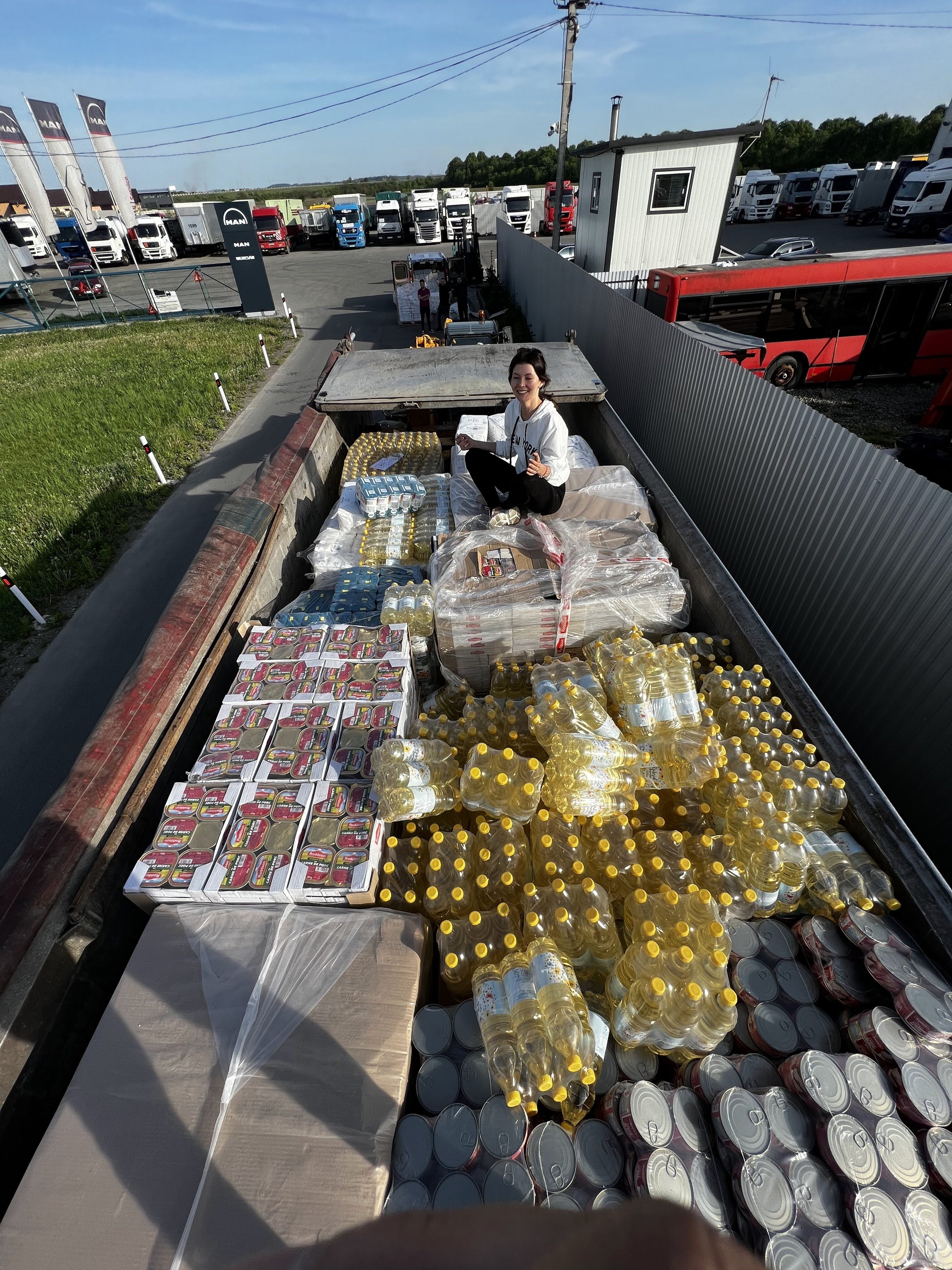 Person sitting in the back of a cargo truck surrounded by bottled water, canned food, and other supplies, with a parking lot full of trucks and a red bus in the background.