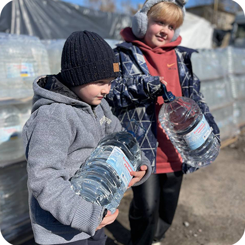 Two children standing outdoors at a recycling center, holding large plastic water bottles.
