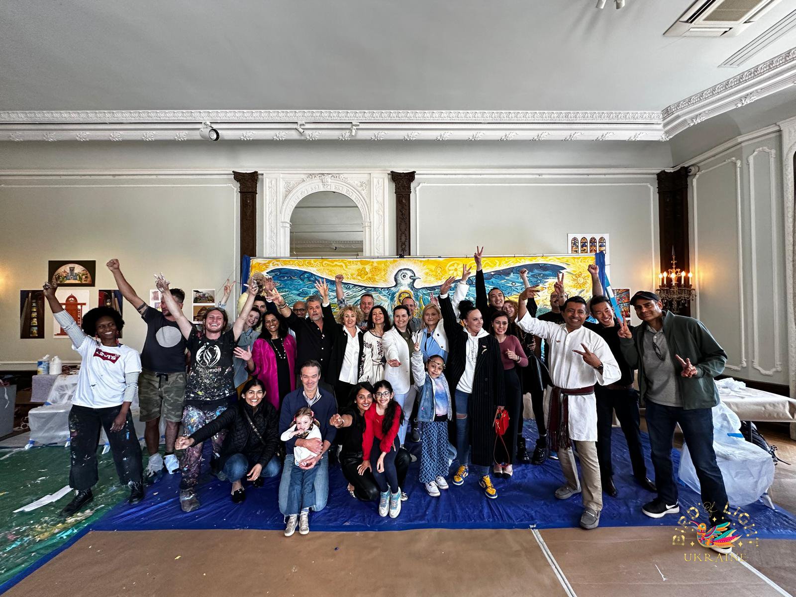 Group of diverse people posing together in front of a large colorful mural of a bird, with some raising their arms and making peace signs, inside a grand room with high ceilings and ornate moldings.