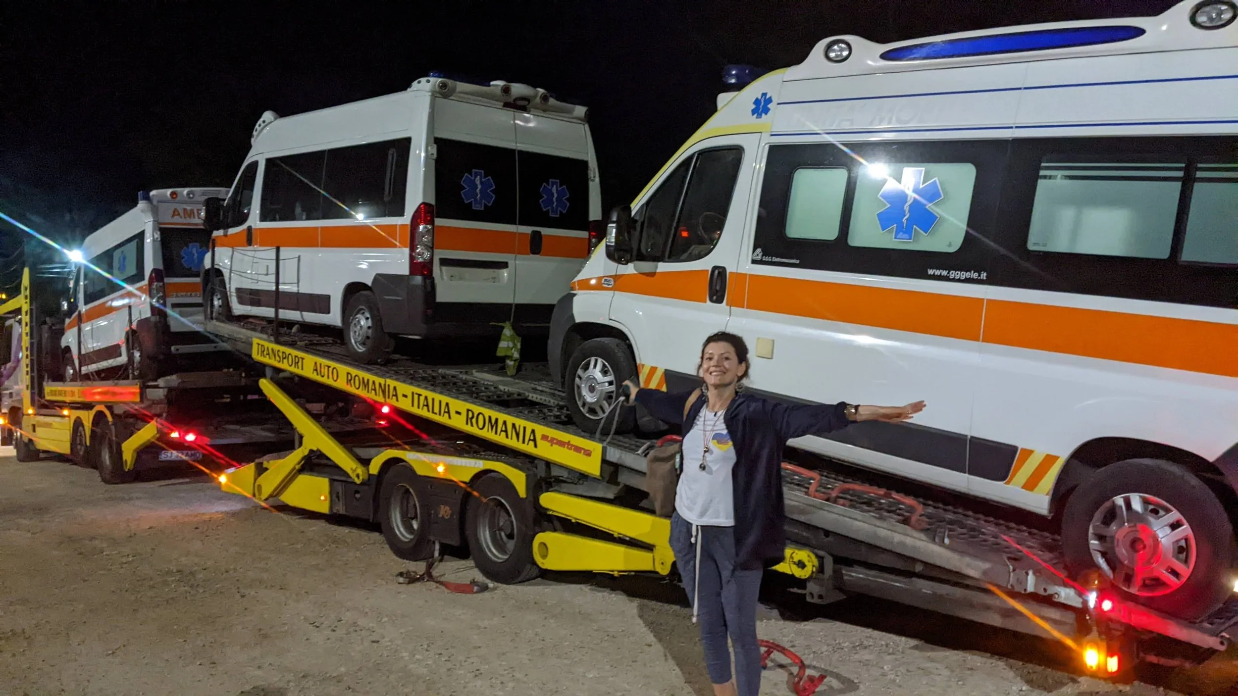 A woman standing in front of a transport truck with three ambulances loaded on it at night. She is smiling and has her arms outstretched.