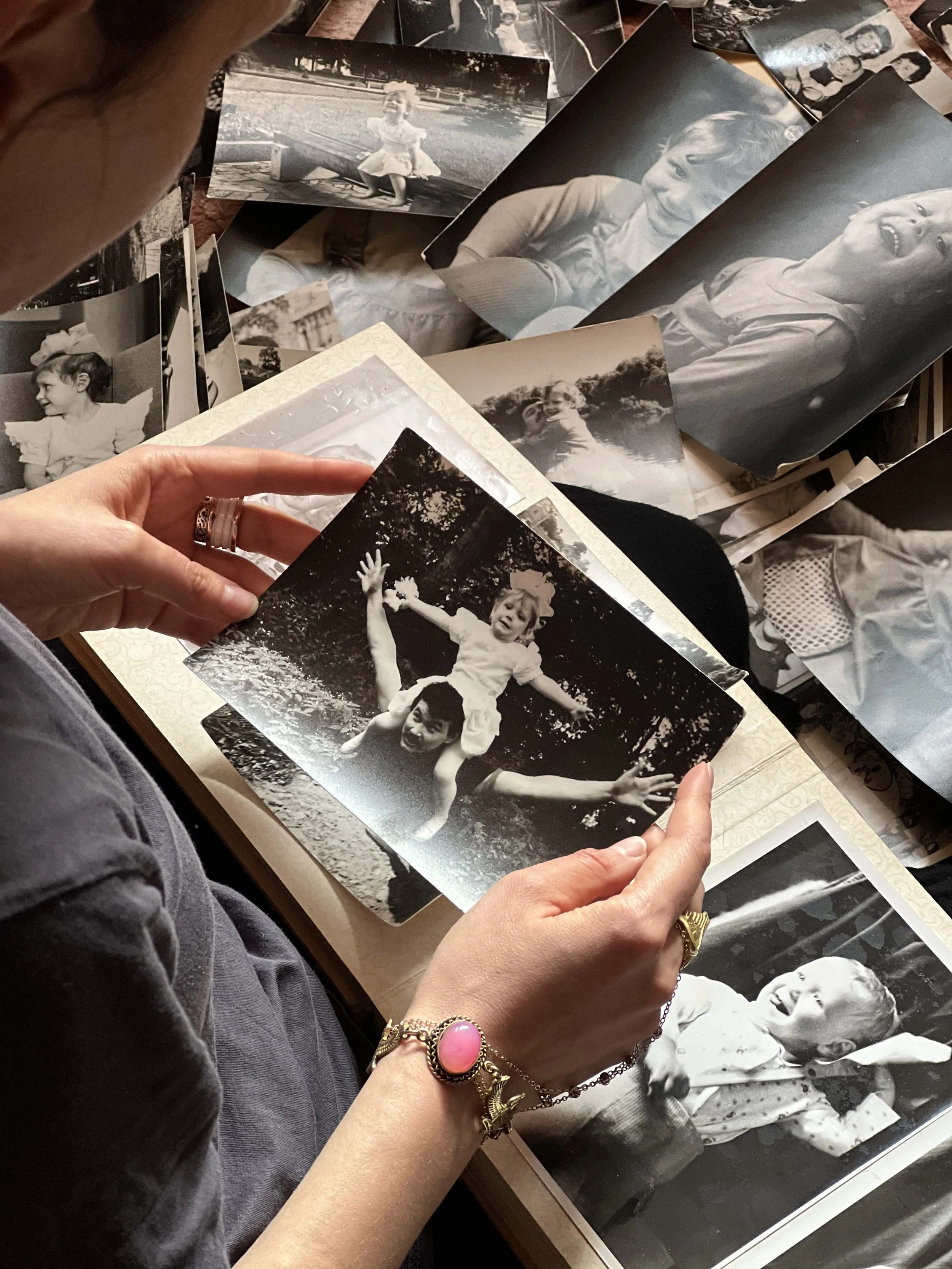 A person holding a black-and-white photograph of a young girl sitting on a man's shoulders, with other black-and-white photos of children scattered on a table.