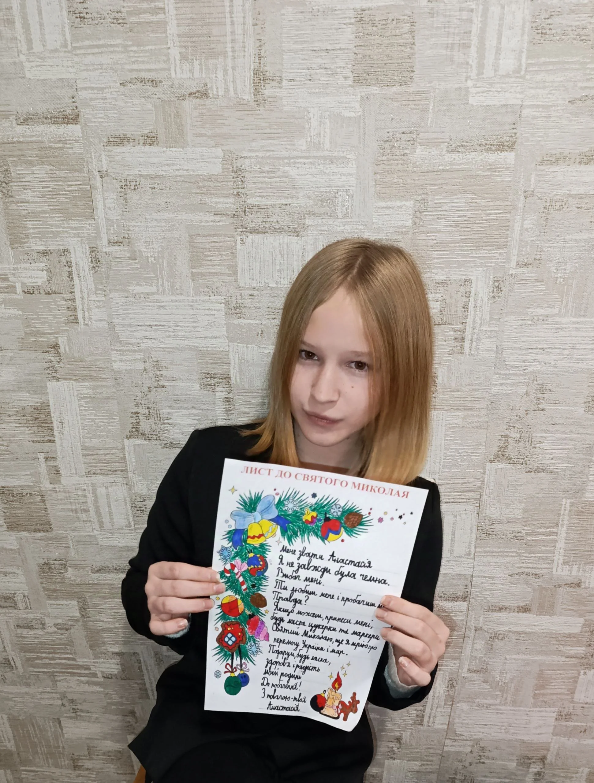A young girl with red hair holding a colorful handwritten letter with Christmas decorations, standing against a textured beige wall.