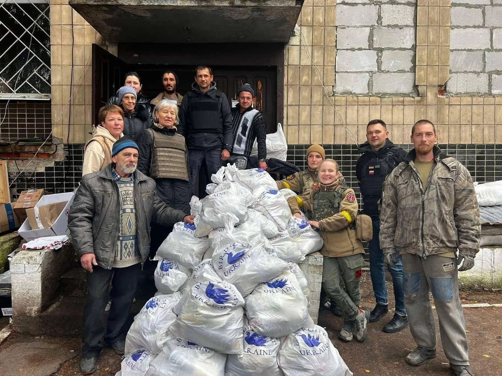 Group of people standing around bags of supplies in front of a partially damaged building, likely involved in aid or relief efforts.
