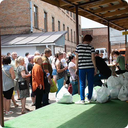 A line of people waiting to receive food from a distribution point outdoors with bags of supplies, under a covered area against a brick building.