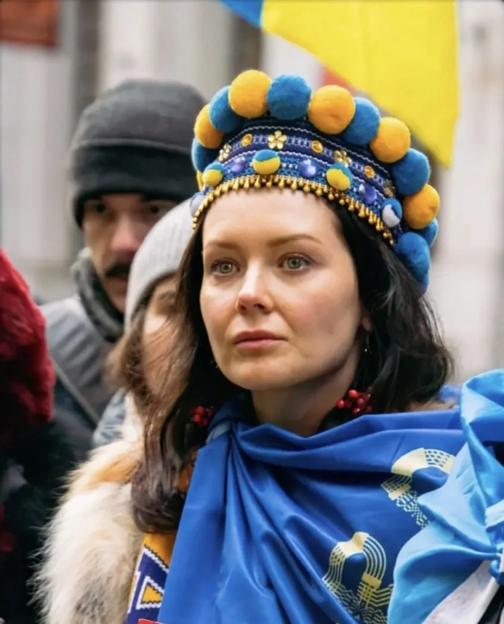 A Ukrainian woman wearing a colorful traditional Ukrainian headdress decorated with blue and yellow pom-poms and embroidered details, standing among a crowd at a cultural or political event.