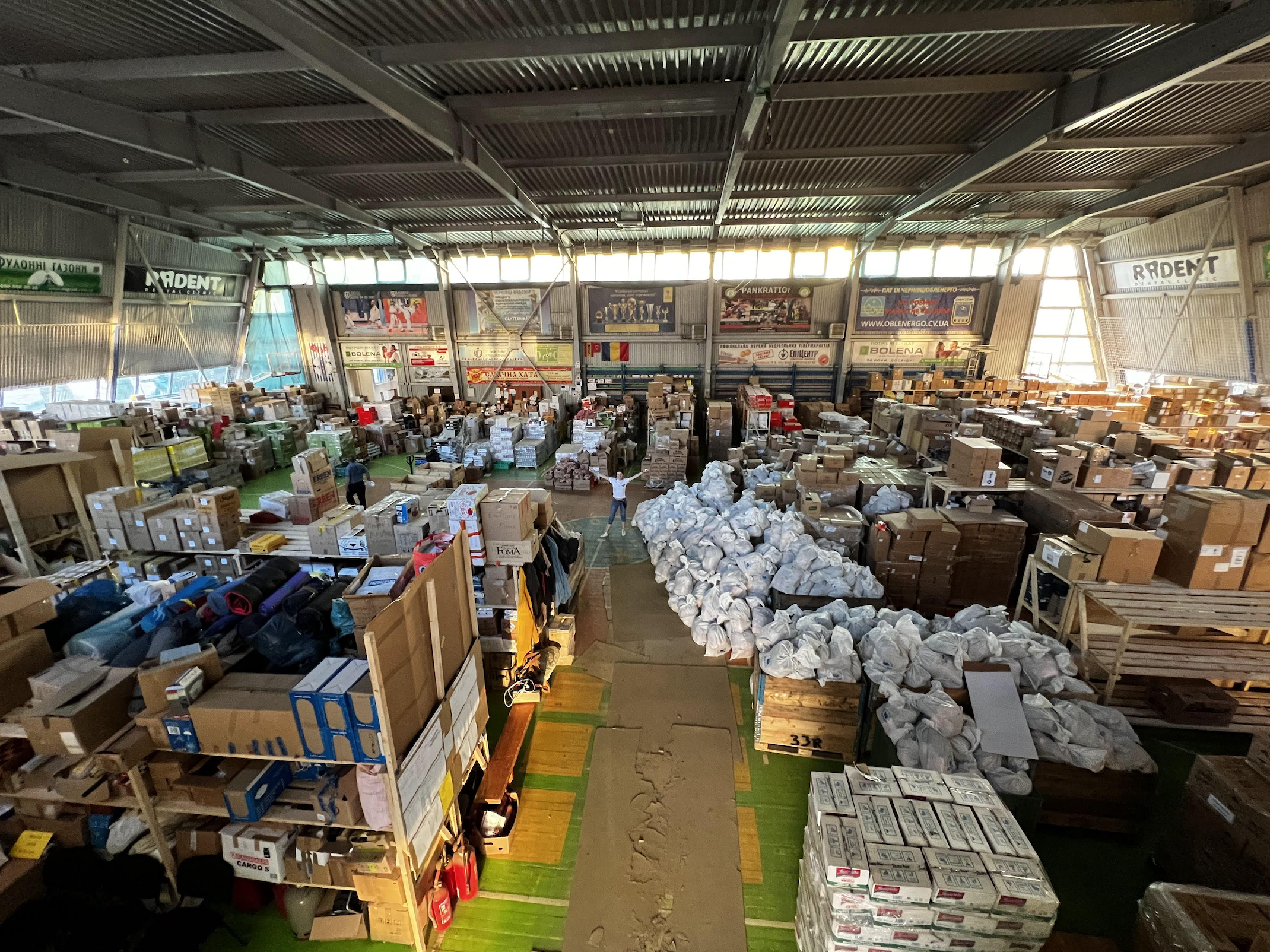 Interior of a large warehouse filled with numerous stacked boxes and packages, with several people working or moving items, and banners hanging from the ceiling.