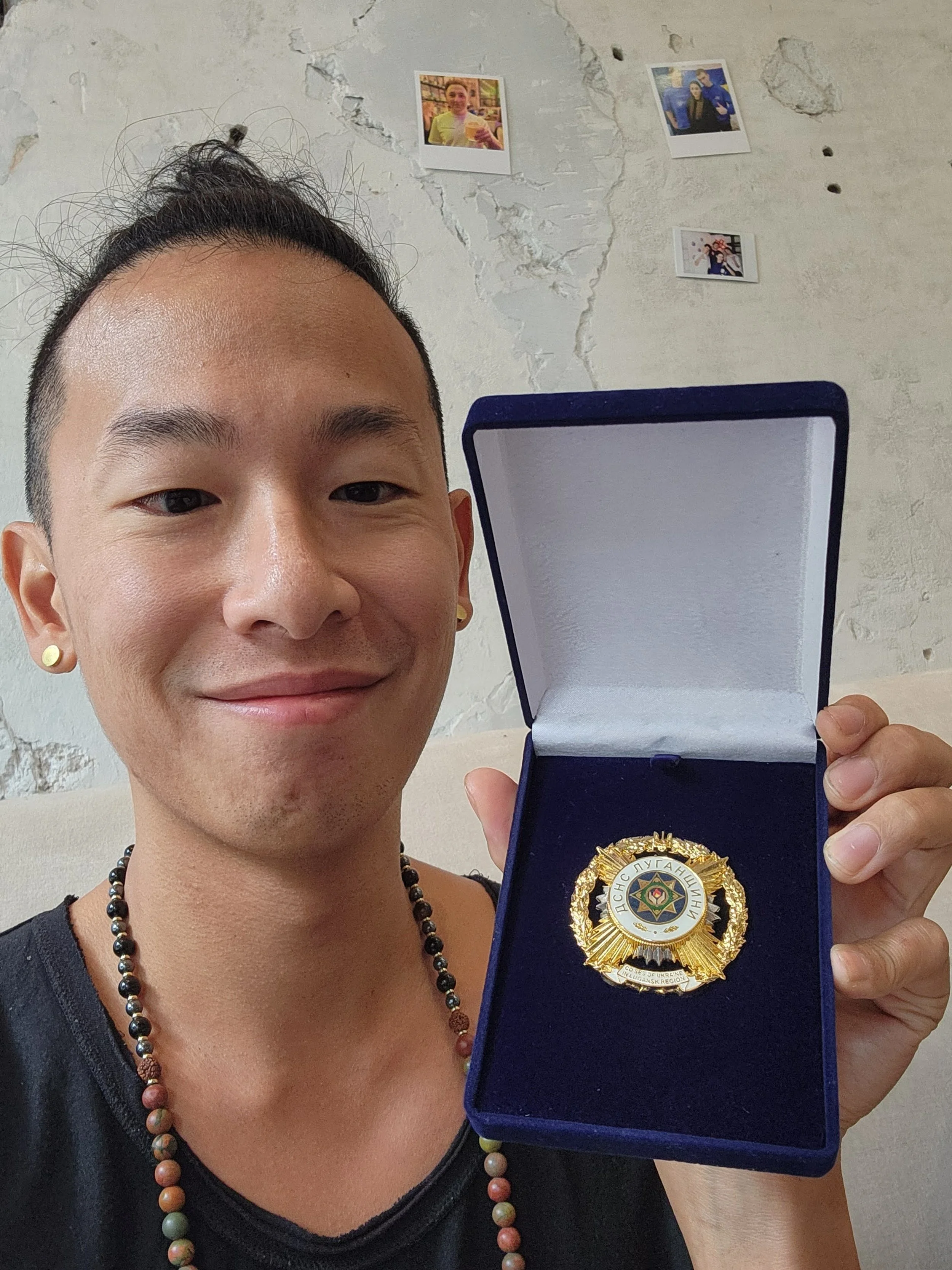 A Chinese Canadian person smiling and holding an award badge in a blue box. The person has short black hair, earrings, and a beaded necklace. The background features a white wall with peeling paint and some small photos taped to it.