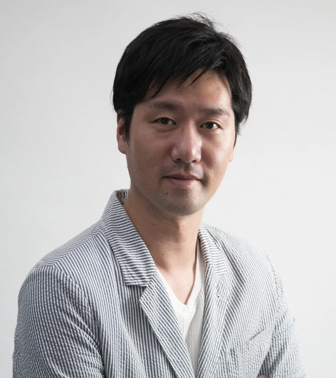 A young Asian man with dark hair, wearing a light-colored blazer over a white shirt, standing with arms crossed against a plain white background.
