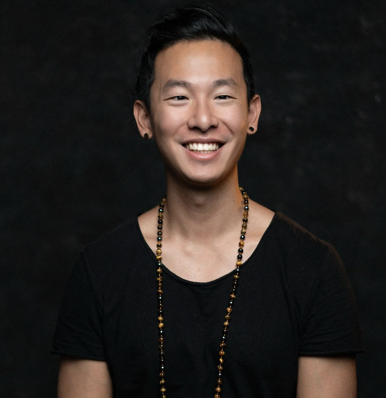 A young man with short black hair, wearing earrings and a beaded necklace, smiling at the camera against a dark background.