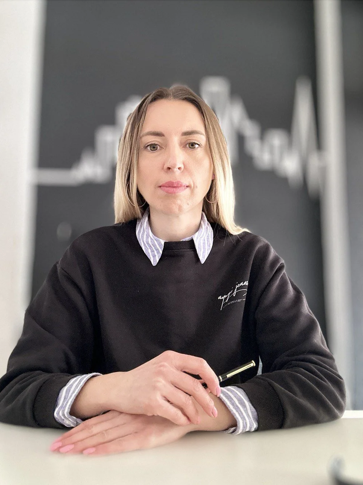 A woman with blonde hair wearing a black sweatshirt and a collared shirt underneath, sitting at a desk with her hands clasped, with a blurred bar chart in the background.