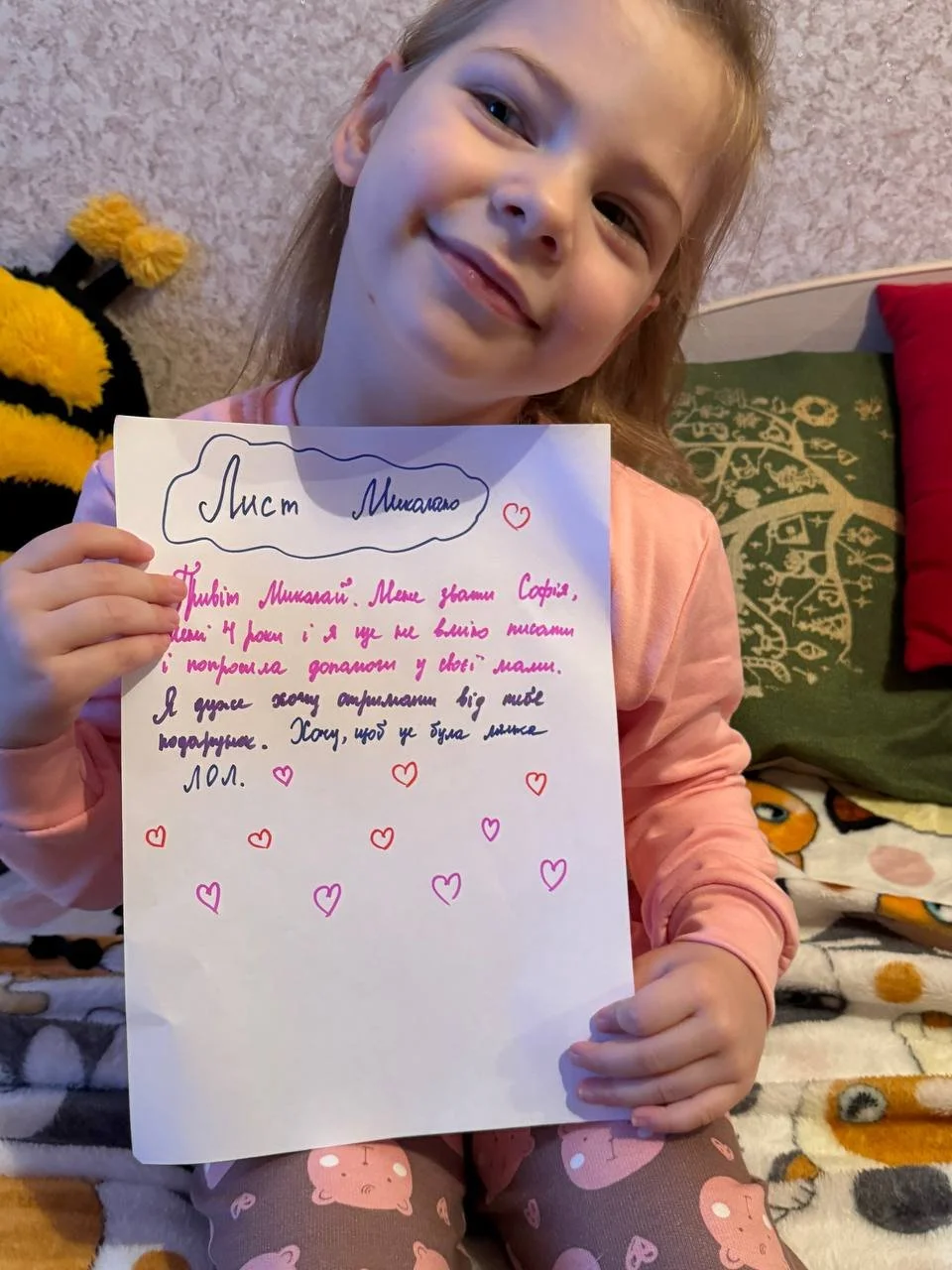 A young girl with a smile holding a handwritten letter with colorful hearts. She is sitting on a bed with a blanket that features cartoon characters, in a room with a textured wall and pillows in the background.