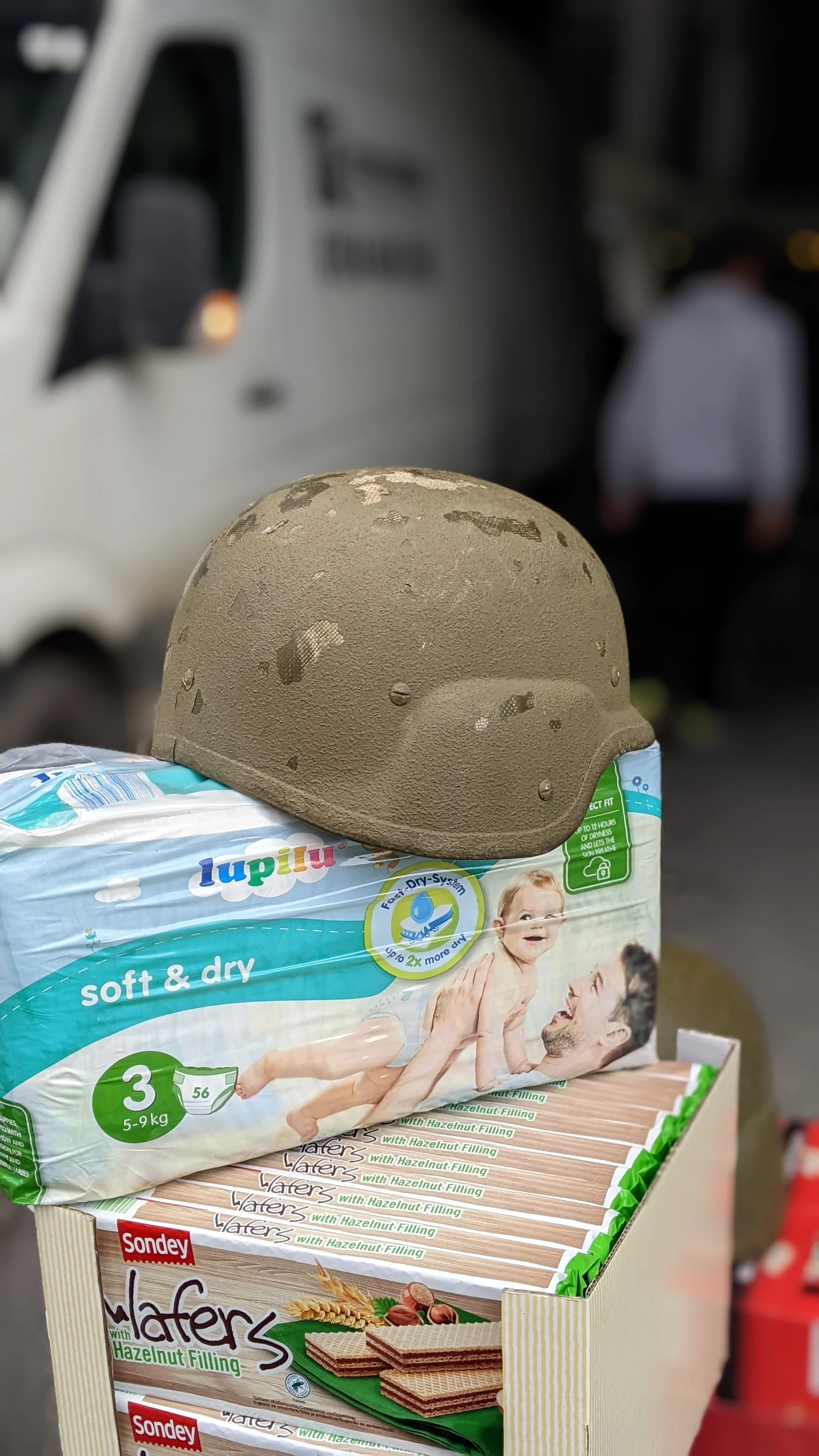 A package of Lupilu soft and dry diapers with a helmet placed on top. Behind the package, a white truck and a person in a white shirt can be seen.