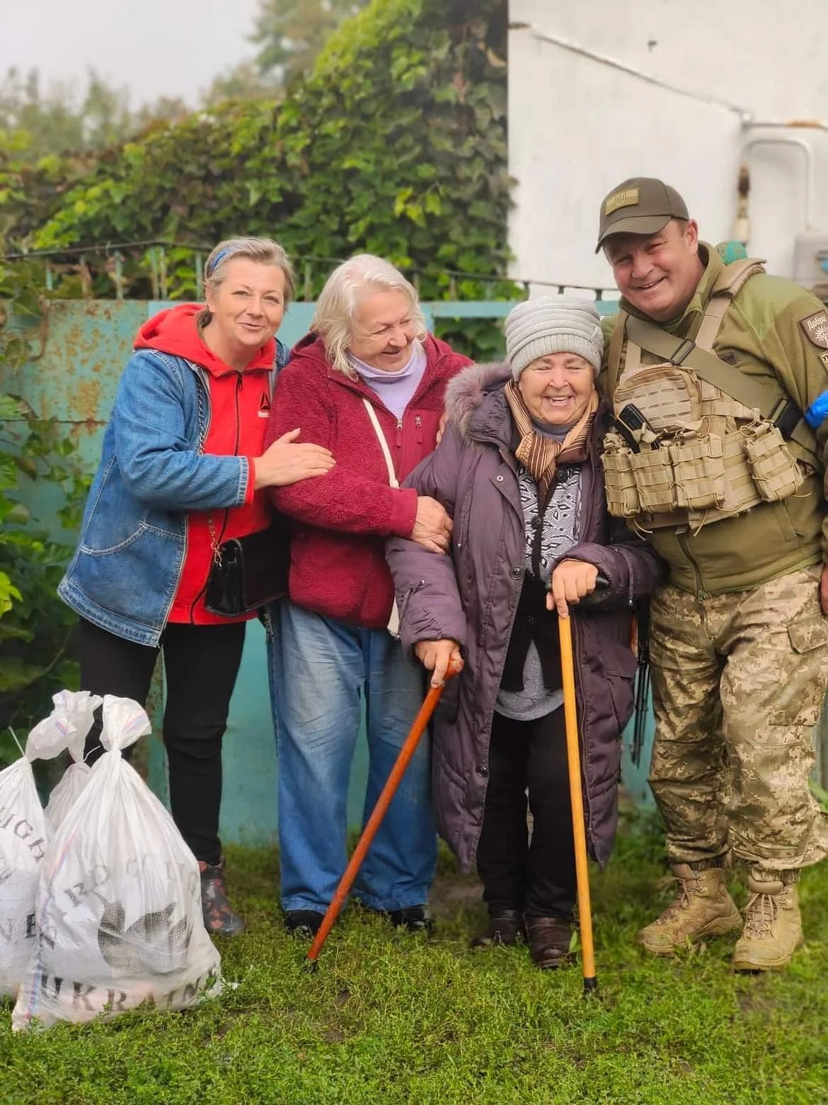 Four Ukrainian women and a Ukrainian man in military uniform standing together outdoors, smiling, with two women using walking sticks. It appears to be a cheerful gathering in a garden or backyard, with bags of humanitarian aid nearby.