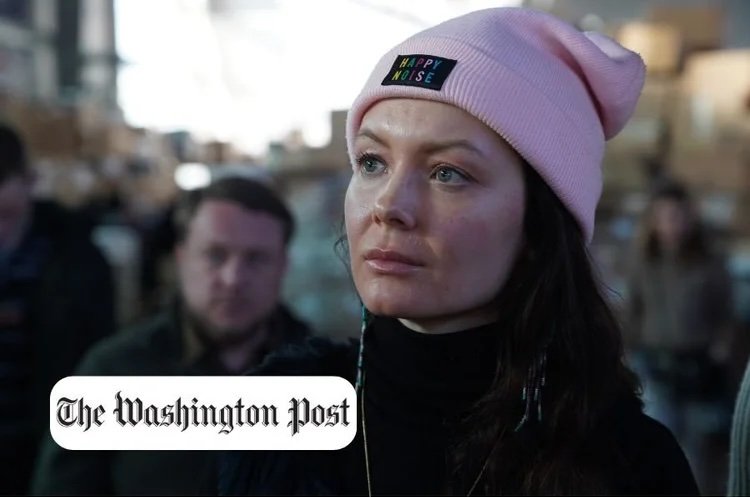 A woman wearing a pink beanie with a 'HAPPY NOISE' logo, looking thoughtfully in an outdoor setting with a blurred crowd in the background.