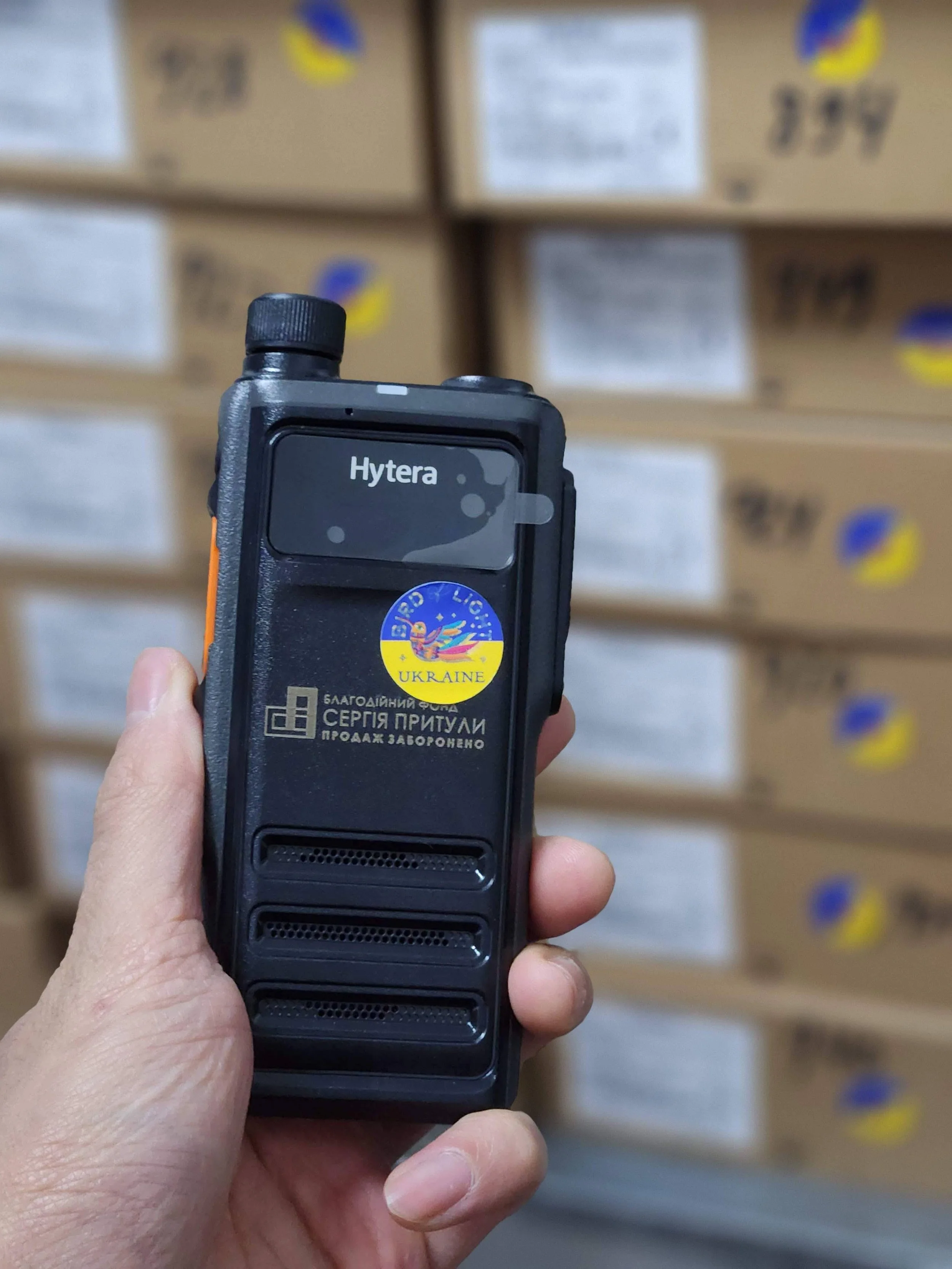 A person holding a black handheld radio device with a sticker that says 'Ukraine' and a logo. The background shows stacked cardboard boxes with labels and logos.