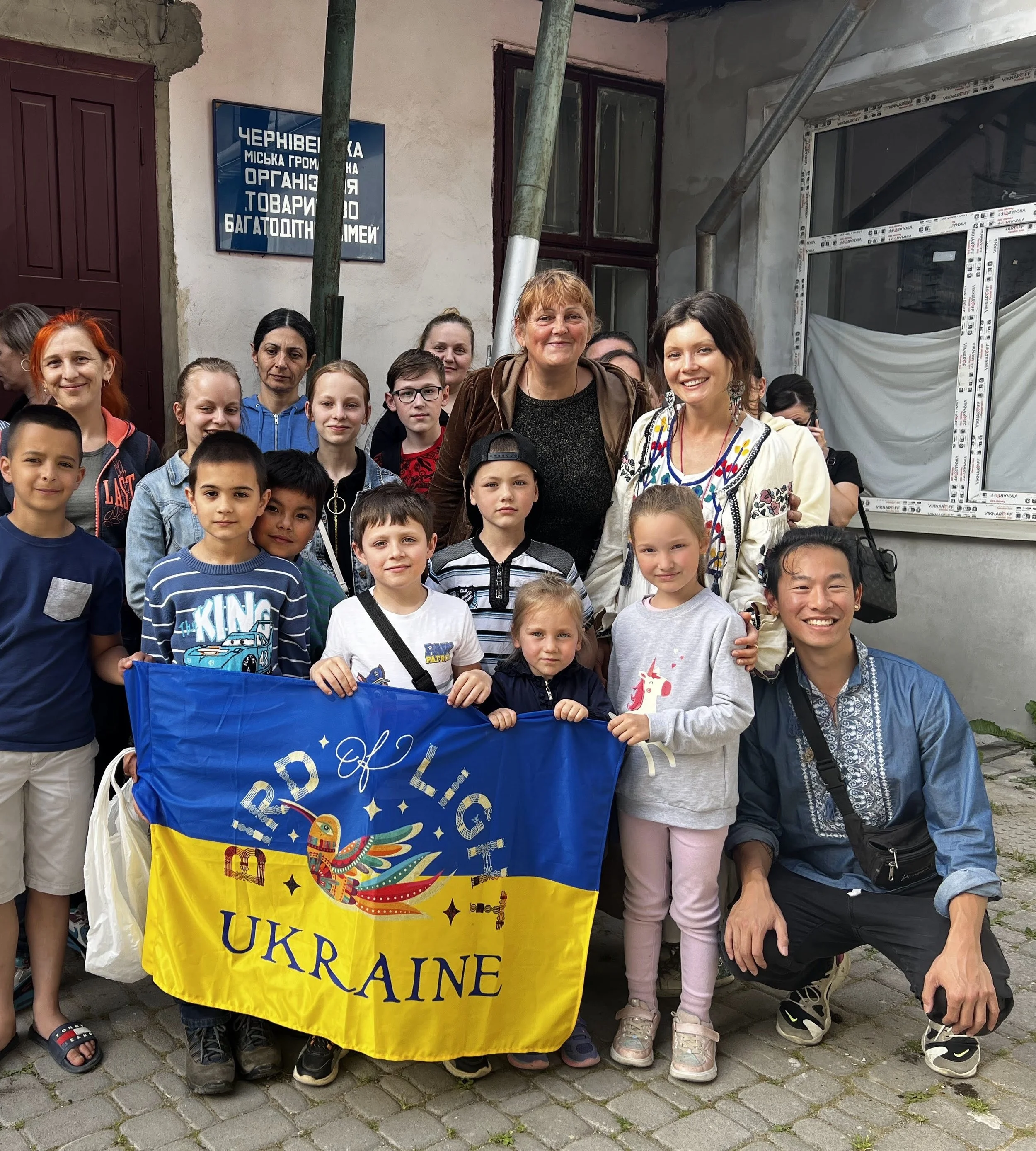 Group of children and adults holding a Ukrainian flag with a colorful bird design, gathered outside a building with a blue sign in Ukrainian, celebrating Ukraine.