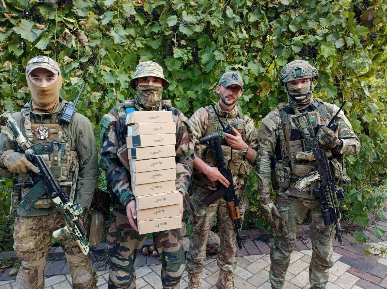 Four military personnel in camouflage uniforms standing in front of a bushy background, holding weapons, with one soldier carrying a stack of boxes.