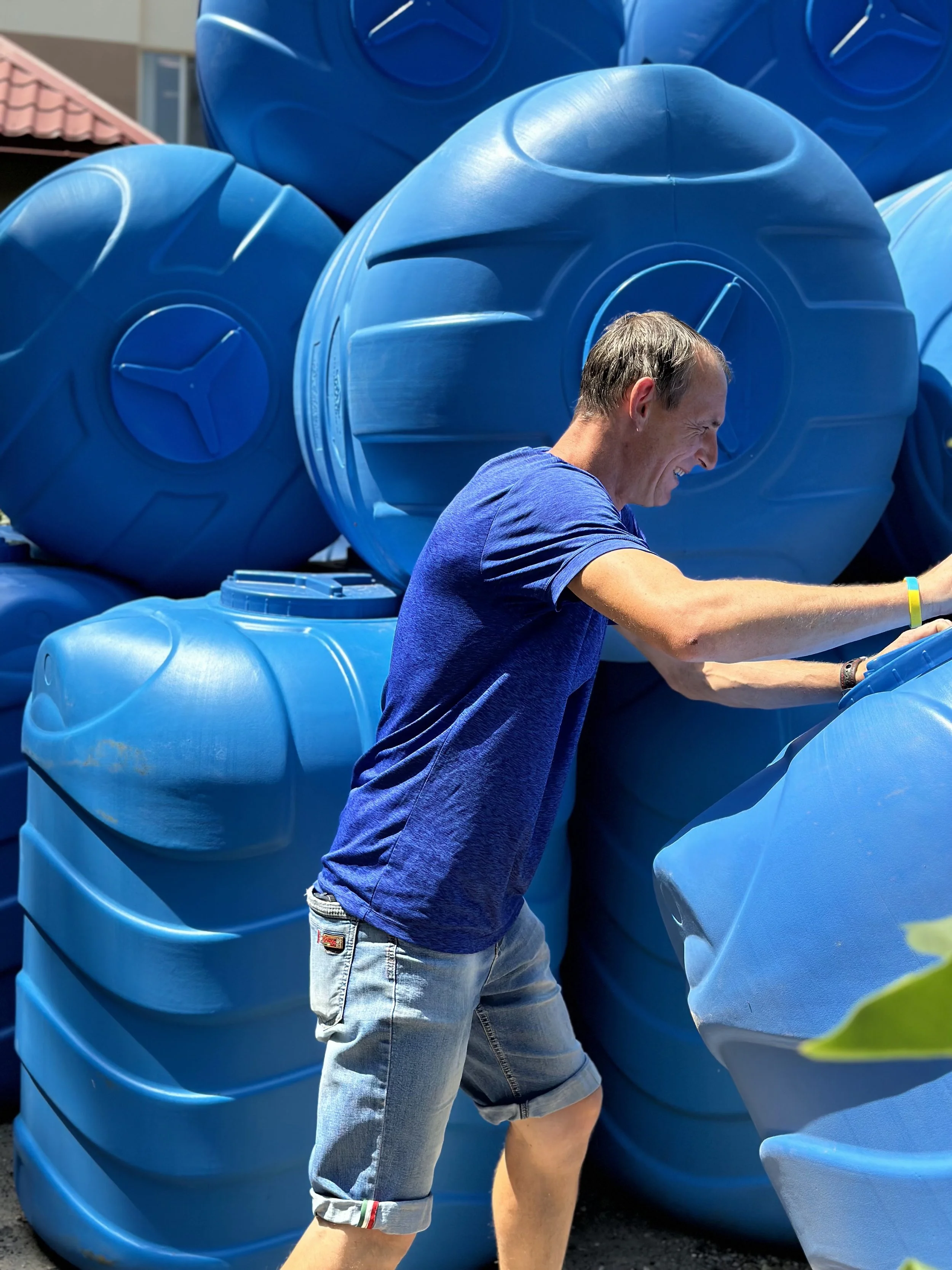 A man in a blue t-shirt and denim shorts smiling and playing among large blue plastic barrels outdoors.