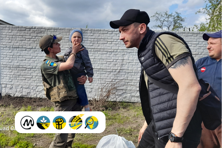 A person in military uniform holding a young child while another man, wearing a black cap and vest, leans forward and interacts with them. Another man stands nearby, smiling. They are outdoors near a white brick wall and some greenery.