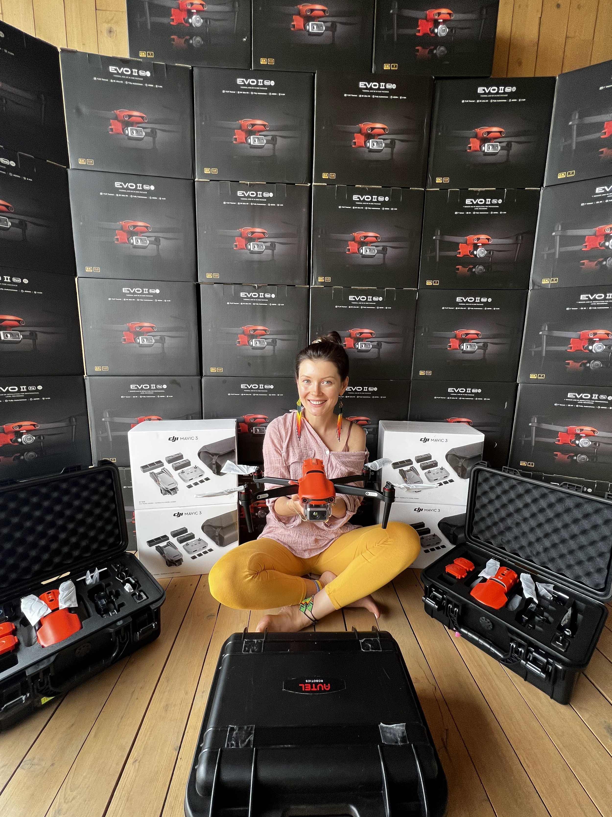A Ukrainian woman sitting cross-legged on a wooden floor, smiling, surrounded by drone equipment and boxes, with a wall of drone boxes behind her.