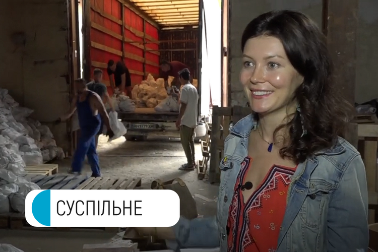 A woman in a denim jacket smiling for the camera inside a warehouse or factory, with workers in the background handling bags or materials
