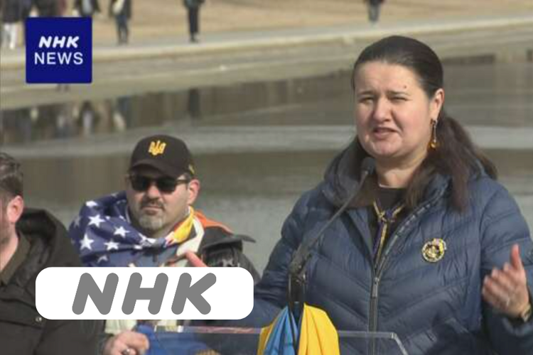 A woman speaking at a public event, with a serious expression, wearing a blue jacket and earrings. Two men are in the background, one wearing sunglasses and a cap with a flag draped over his shoulders. There is a logo in the top left corner that says 'NHK NEWS'.